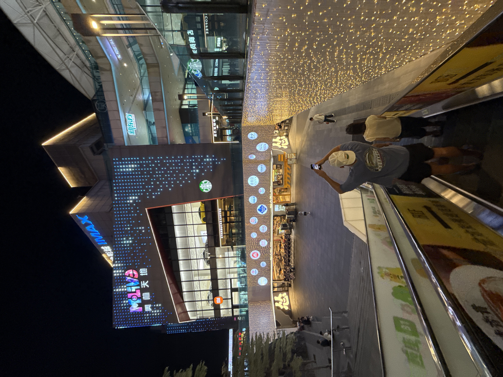 The image captures a night scene at a bustling shopping mall in Tianhe District, China. The architecture is modern and brightly lit, with illuminated signage such as "MOLIVE," "IMAX," and "Starbucks Coffee." The facade of the building is adorned with a vibrant pattern of blue and white lights. Below the main facade, a lower level is decorated with numerous small, golden lights, creating a festive atmosphere.
In the foreground, two individuals are visible on an escalator, with one person taking a photograph of the scene with their phone. Several other people are scattered throughout the plaza area, some walking, others seated, suggesting a lively evening. The plaza itself features various shops and restaurants, and the overall impression is one of urban vibrancy and commercial activity. The backdrop of the dark night sky emphasizes the illuminated nature of the shopping complex.