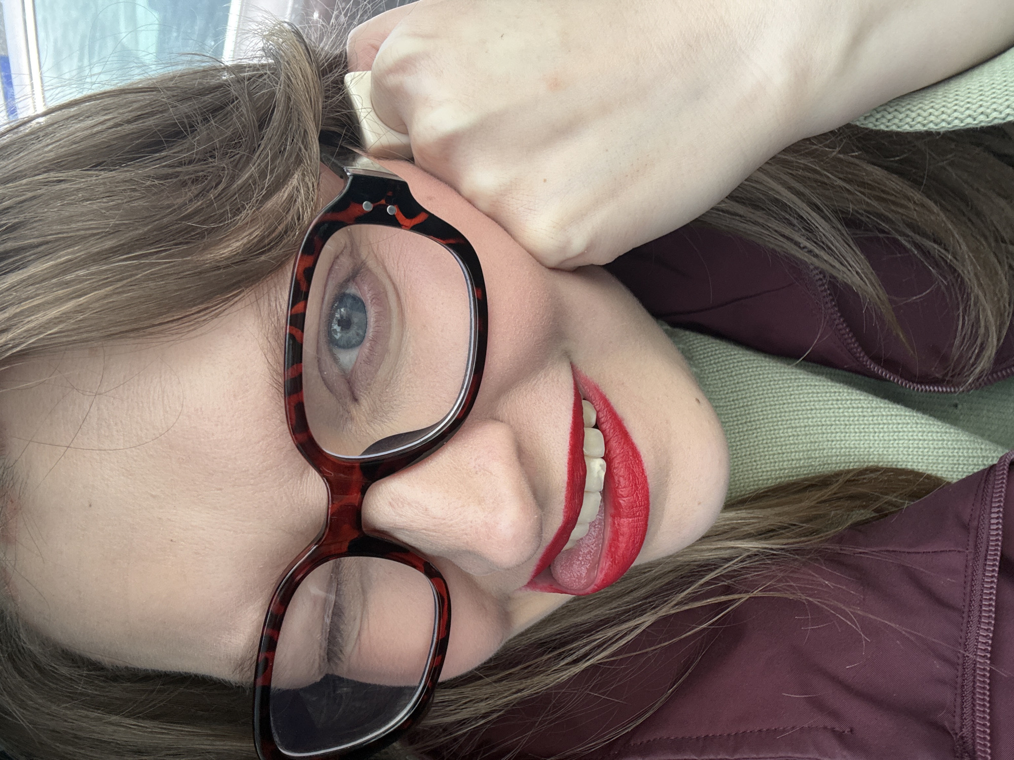 A woman with fair skin, wearing red lipstick and tortoise-shell glasses, is seen in a close-up shot. She has light brown hair. She is holding her hand against her face. She is wearing a maroon jacket and a green knit sweater. It is not possible to determine the location of the image, nor whether the location is in Izhevsk, Russia.