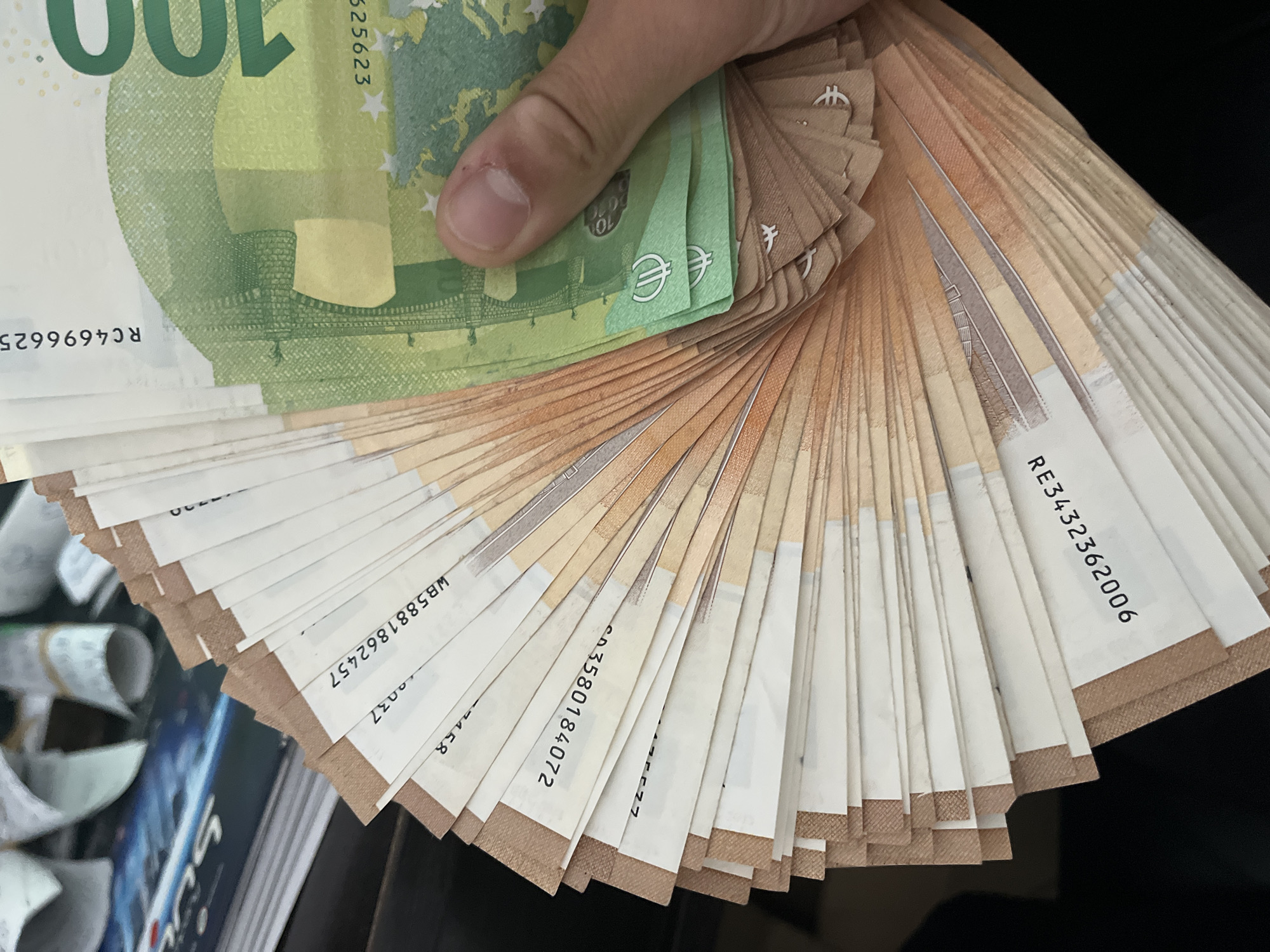 A person's hand is prominently displayed, holding a large fan of Euro banknotes against a dark, indistinct background. The hand's thumb and fingers are visible, gripping the currency at the top.
The top notes in the fan are green 100 Euro banknotes, with the number "100" clearly visible. Below these, a large quantity of orange-brown 50 Euro banknotes are fanned out, creating a layered effect. Under the thumb, several smaller green notes, likely 10 Euro denominations, can be seen with a "10" and a Euro symbol (€).
Visible text on the 100 Euro notes includes the serial numbers "RC4696625" and "025623". European stars and a partial map of Europe are also discernible. On the fanned 50 Euro notes, multiple serial numbers are visible, including "WB5881862457", "CD3580184072", and "RE3432362006", along with other partial numbers like "0037" and "458". Several Euro symbols (€) are also seen on the visible notes.
The scene appears to be indoors, under artificial lighting. The background is largely out of focus, with blurry shapes suggesting other objects or surfaces to the left and dark space to the right. The image captures a moment of displaying a significant amount of cash, located in Ivailo, Bulgaria.
