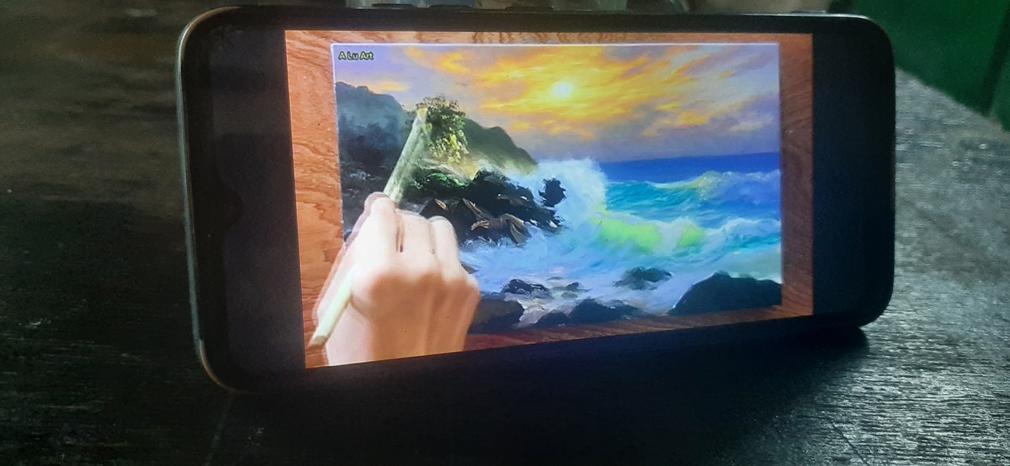 A horizontal, eye-level, medium shot captures a smartphone displaying a digital painting of a seascape. The painting depicts a dramatic scene with crashing waves against rocky cliffs under a vibrant sunset sky. A hand is shown reaching into the frame from the left, holding a paintbrush, as if digitally painting the scene. The smartphone is placed on a dark, textured surface, possibly a table, with a blurred green background visible behind it. The lighting is dim, with the glow of the smartphone screen illuminating the immediate surroundings. The artwork on the screen is signed "ALu Art" in the upper left corner. The image does not contain any information about the city of Yessentukskaya, Russia.