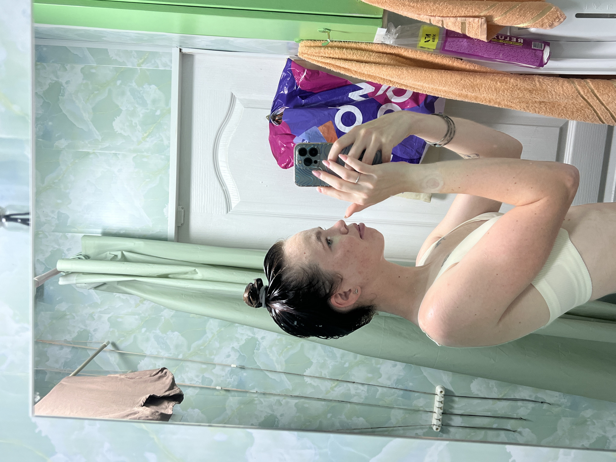 In the city of Chaikovsky, Russia, a young woman stands in what appears to be a bathroom, taking a selfie in a mirror. She has dark hair pulled up in a bun and is wearing a light-colored bra. Her skin is speckled with freckles, and she has a faint tan line on her right arm. The background of the mirror reflects a bathroom with light green marble-patterned walls, a mint green shower curtain, and a white door. Various items are visible in the reflection, including a towel rack with a brown towel, a purple shopping bag, and some cleaning supplies. The overall scene suggests a private, domestic setting.