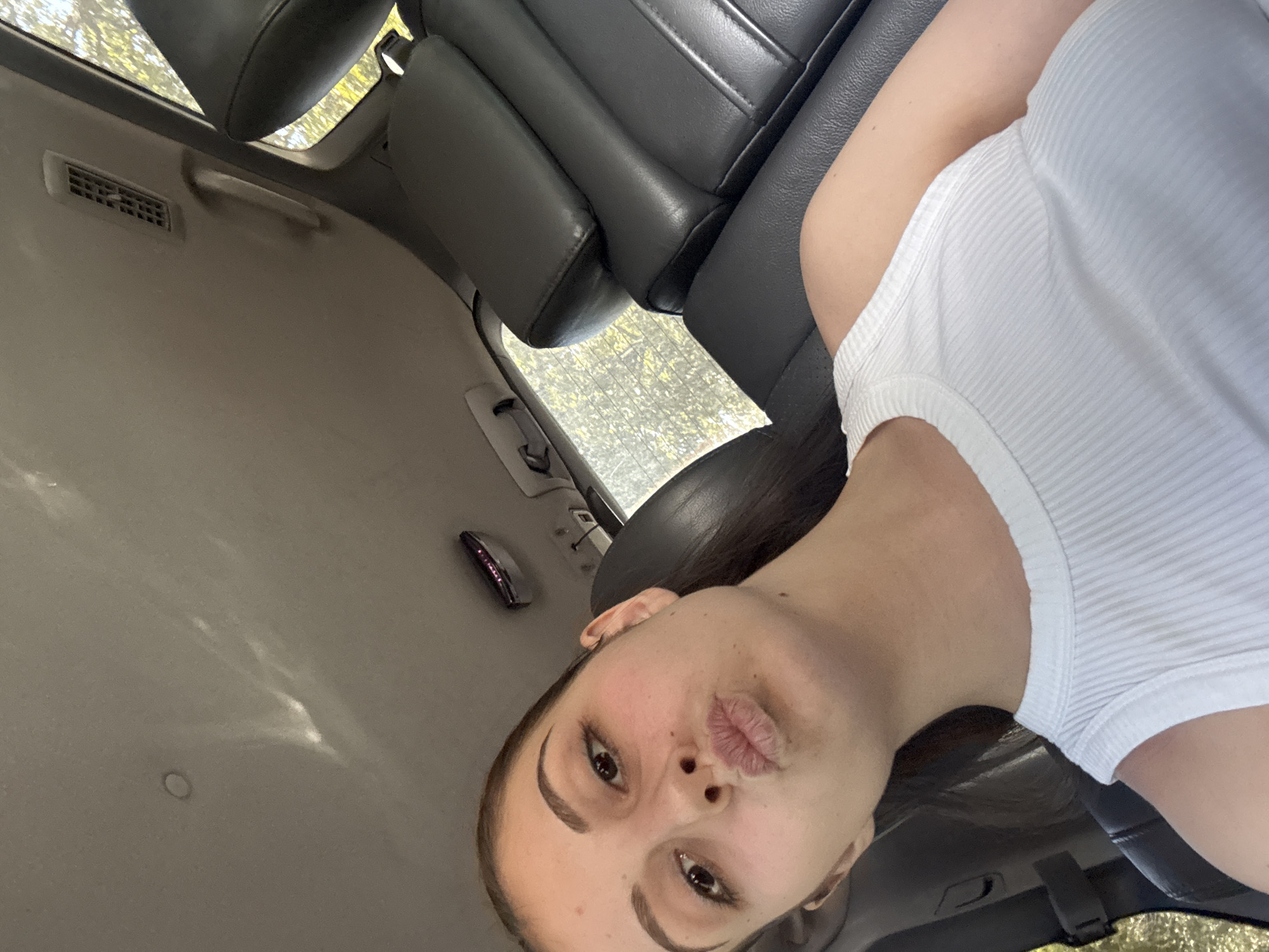 The image, taken inside a car, features a young woman taking a selfie during bright daylight. Her face is prominently displayed, angled slightly upwards, with dark hair pulled back from her face. She has dark, neatly shaped eyebrows and is making a playful or "pouty" expression with her lips. She is wearing a white, ribbed tank top, visible on her right shoulder and chest.
The background shows the interior of a vehicle. Black leather seats are visible to her left and behind her. The car's light-colored headliner (ceiling) is seen above and to her left, equipped with a rectangular air vent and a grab handle. Further back on the headliner, a small, dark, and possibly reflective component (such as a dome light or glasses holder) is mounted. Through the car windows, blurred green foliage and bright sunlight indicate an outdoor, sunny environment. No text is visible in the image. The setting is a car in Burgas, Bulgaria.