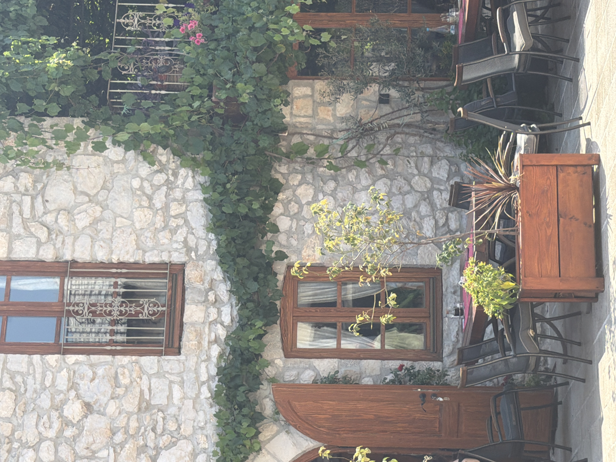 The image depicts an outdoor seating area of a restaurant with a stone building facade, located in Trebinje, Bosnia and Herzegovina. Ivy vines drape over the facade and a balcony. Several wooden chairs and tables are arranged on a tiled patio. The building features a large wooden door and multiple windows with wooden frames and iron security bars. A wooden planter box with green foliage sits in the foreground. The scene is bathed in sunlight, suggesting daytime.