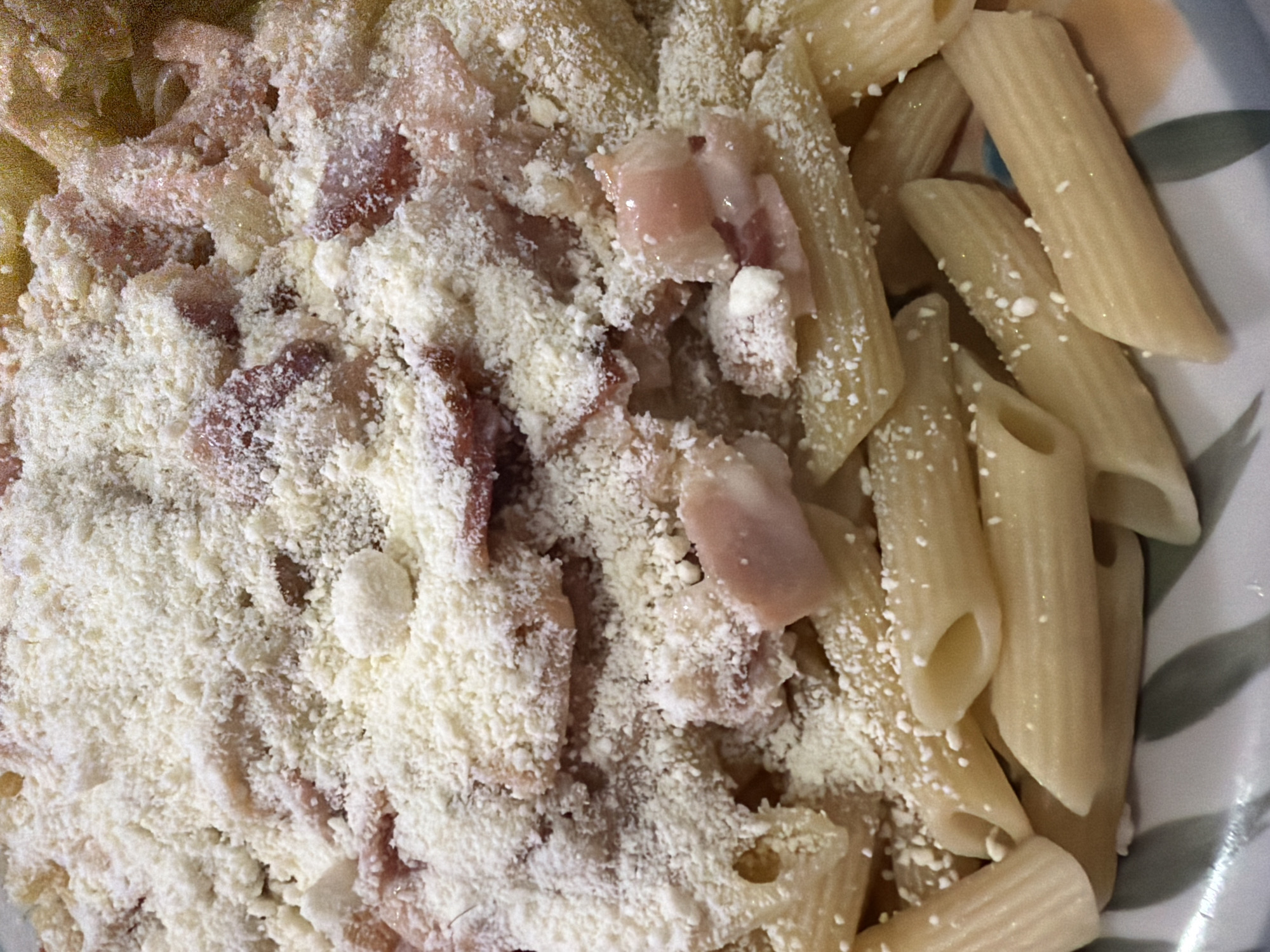 A close-up photograph shows a portion of pasta, likely penne, served in a bowl. The pasta is mixed with chopped meat, possibly ham or bacon, and covered with a generous amount of grated cheese. There are no people visible in the image. The exact location within the city of Ciudad Guayana, Venezuela cannot be determined from the image.