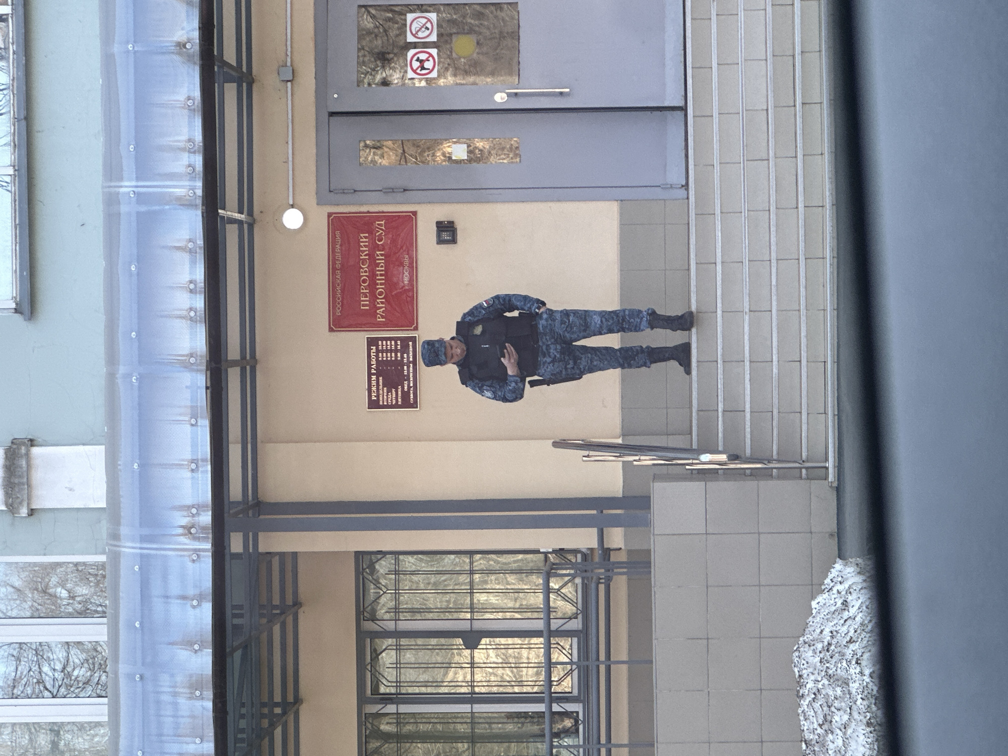 A uniformed security officer stands on the steps leading to the entrance of the Perovsky District Court in Moscow, Russia. The building exterior is beige with gray doors and a sign indicating the court's name and jurisdictional area. A schedule of operating hours is posted next to the entrance. A protective canopy covers the entrance area. The image appears to have been taken from inside a vehicle.