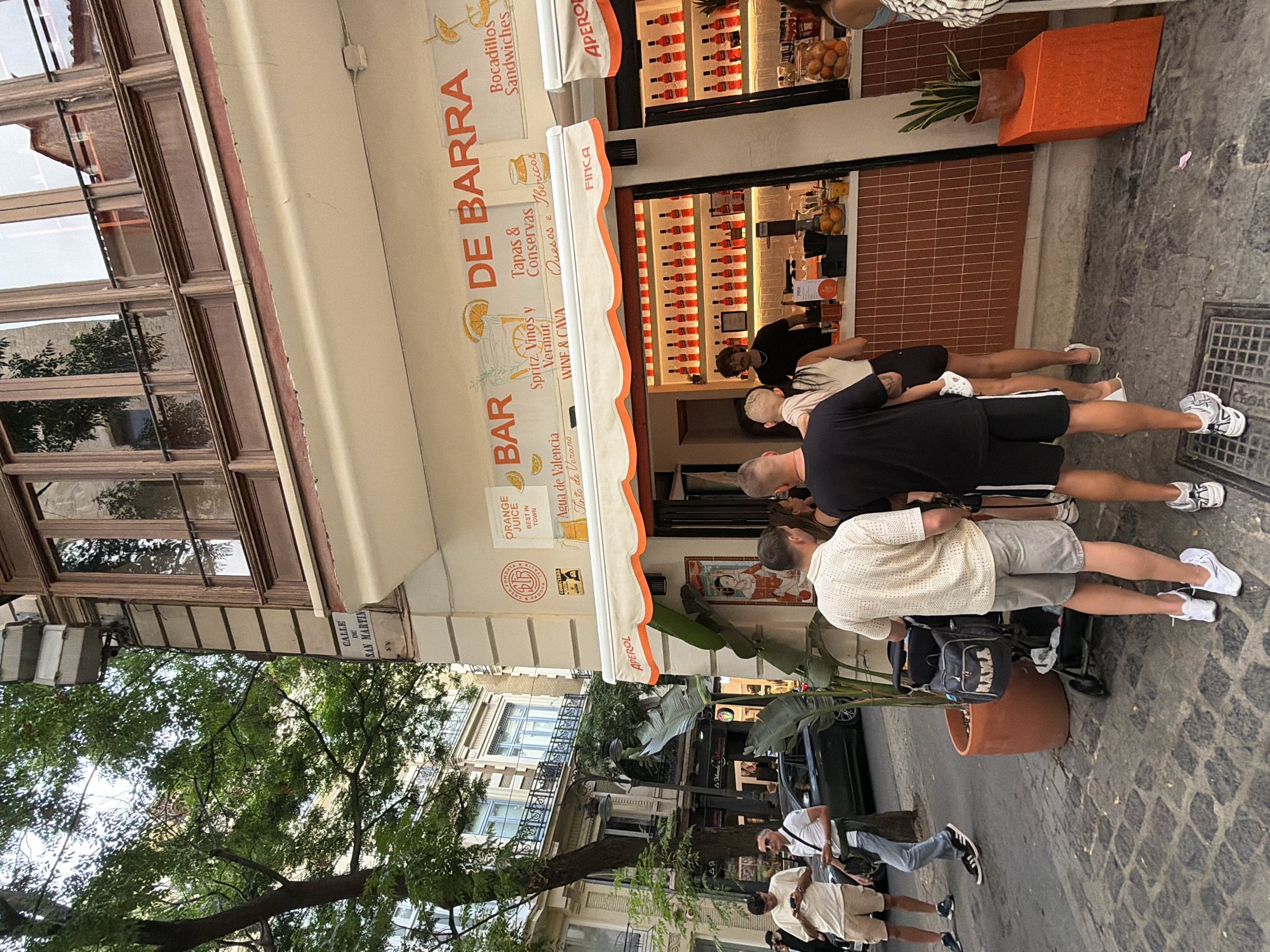 This image depicts a street scene outside a bar with an outdoor service window. In the foreground, several people are standing, appearing to be waiting to order. Two men are facing away from the camera, one wearing a white patterned shirt and grey shorts, the other in a black t-shirt and black shorts, holding a child. Another man in a white polo shirt and sunglasses is visible to the left, looking at his phone. Behind them, a dark-colored car is parked on the street, and a tree with dense green foliage provides shade. The bar itself has an orange and white awning and a counter where drinks are being served. Bottles of alcohol are displayed on shelves behind the counter, and various citrus fruits are visible. A potted plant sits to the right of the service window. The street is paved with cobblestones. The architecture suggests an urban European setting, with a building facade featuring balconies and windows visible in the background. A street sign reads "CALLE DE SAN MARTIN".