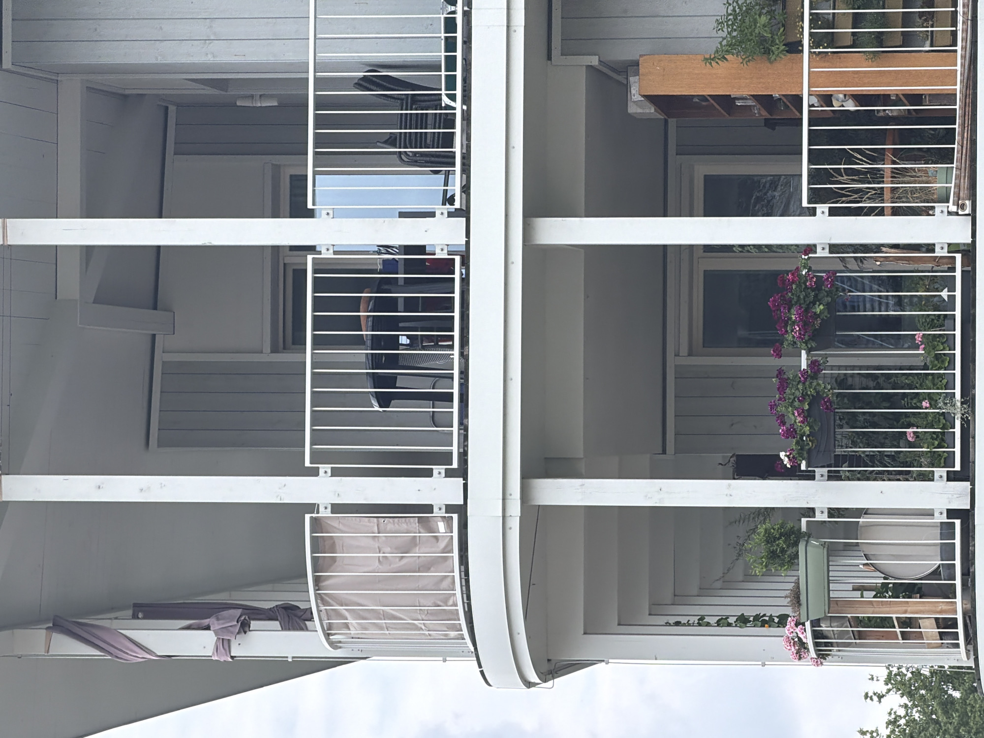 This is a vertical, medium-shot, outdoor photograph of balconies on a modern building, likely an apartment complex. The balconies are constructed with white metal railings. The building's exterior walls are painted a light, muted grey. On the balconies, there are various potted plants, some with vibrant pink and purple flowers, adding splashes of color to the scene. Some balconies have outdoor furniture, including what appears to be a dark chair with a mesh seat and back. There are also some items that look like storage crates or shelves made of wood. On one balcony, a clothes drying rack with a light-colored fabric cover is visible. Some fabric items, possibly clothes or linens, are draped over a railing on a lower balcony. The overall setting suggests a residential area. The sky is visible at the top and bottom of the image, indicating it's daytime, and the lighting suggests it is not directly sunny, perhaps overcast or late afternoon. There is no visible text in the image. The scene conveys a sense of everyday life and domesticity.