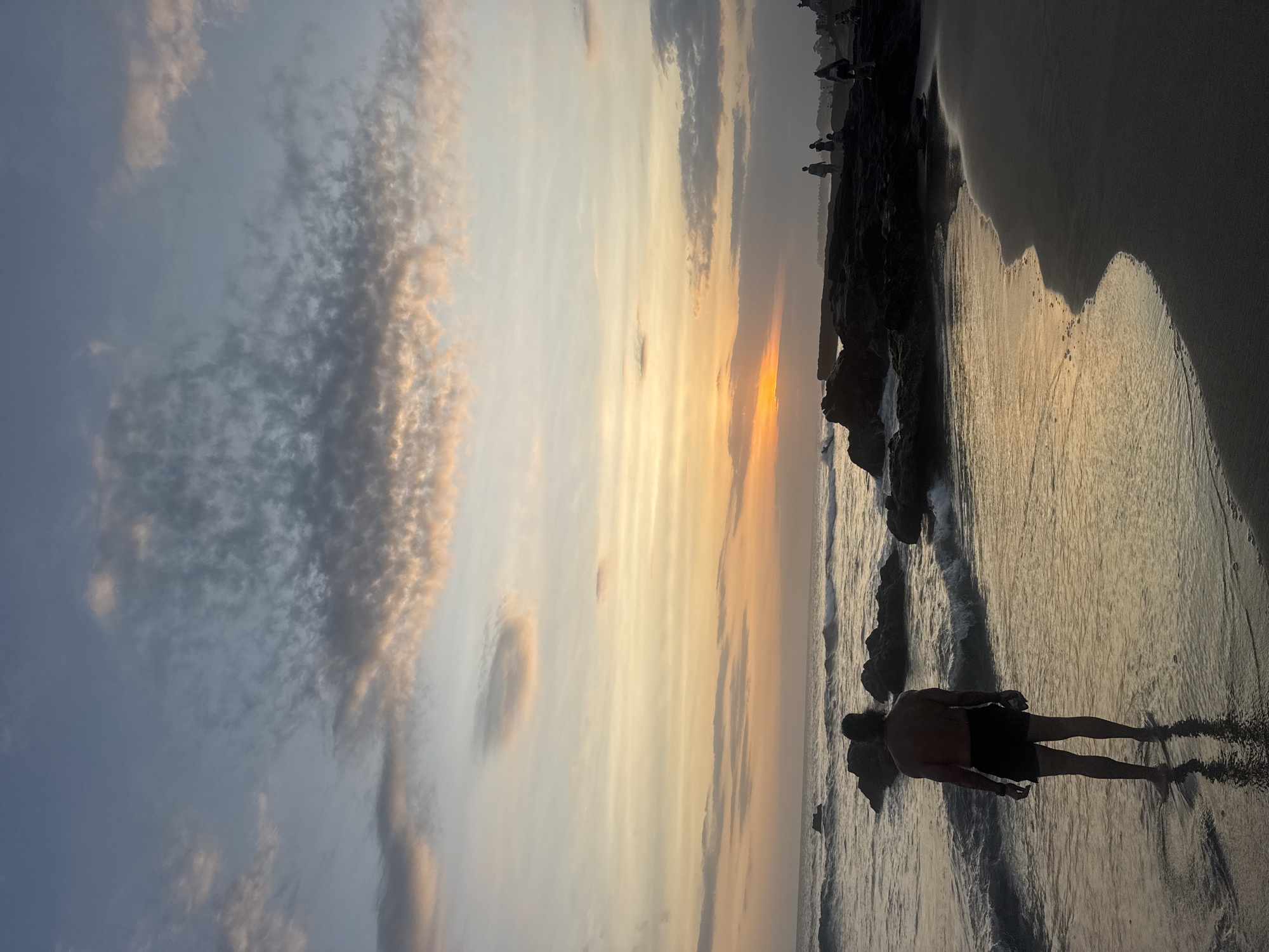 This is a vertical, silhouetted photograph taken at sunset on a beach in North Kuta, Indonesia. The sky is a canvas of warm oranges, yellows, and soft grays, with scattered clouds reflecting the fading light. The ocean meets the sandy shore, with gentle waves lapping at the edge.
In the foreground, a lone male figure stands partially in the shallow water, his silhouette stark against the bright ocean and sand. He is facing away from the camera, looking out towards the horizon. The wet sand reflects his form, creating a mirror image. Further down the beach, other silhouetted figures can be seen walking along the shore, adding to the serene atmosphere.
The setting is clearly a beach, with the characteristic sand and sea. The sunset indicates it's late in the day, likely early evening. The weather appears clear with some cloud cover, typical for a tropical sunset. The overall mood is one of peace and tranquility, inviting contemplation of nature's beauty. There is no visible text in the image.