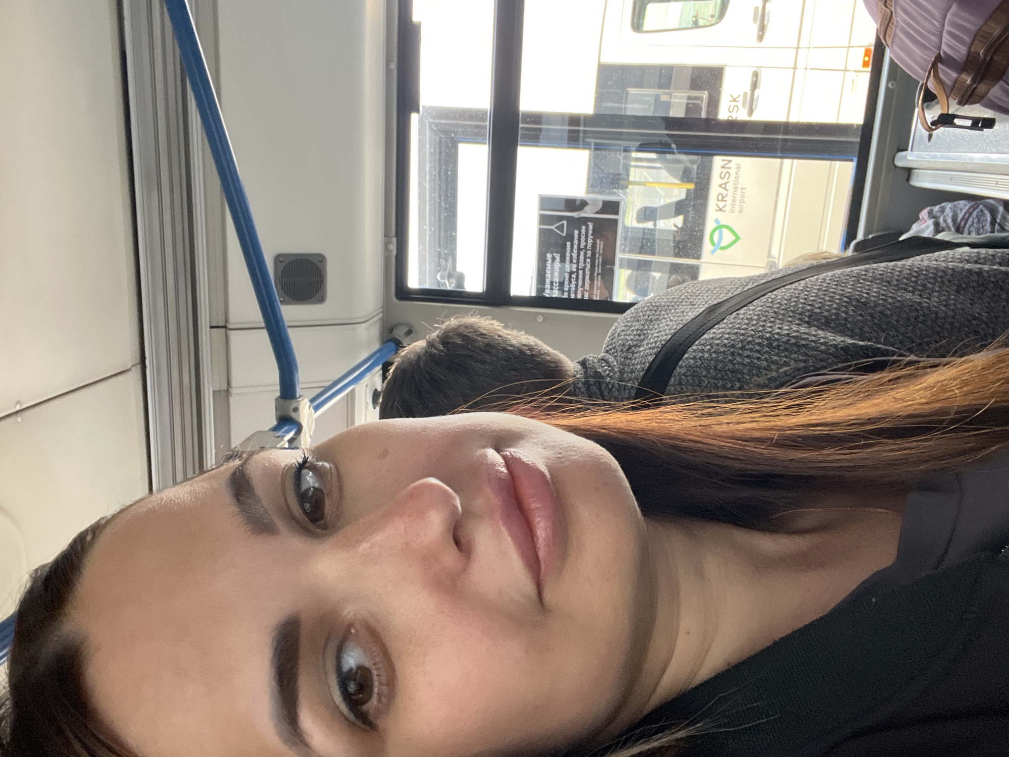 A woman is taking a selfie on a bus. She is in the foreground, her face taking up most of the left side of the frame. She has dark hair, brown eyes, and is wearing light pink lipstick. She is looking at the camera with a slight smile. Behind her, the interior of the bus is visible, including blue handrails and a window. Through the window, another bus is visible with the text "KRASN international airport" on it. The overall lighting suggests it is daytime.