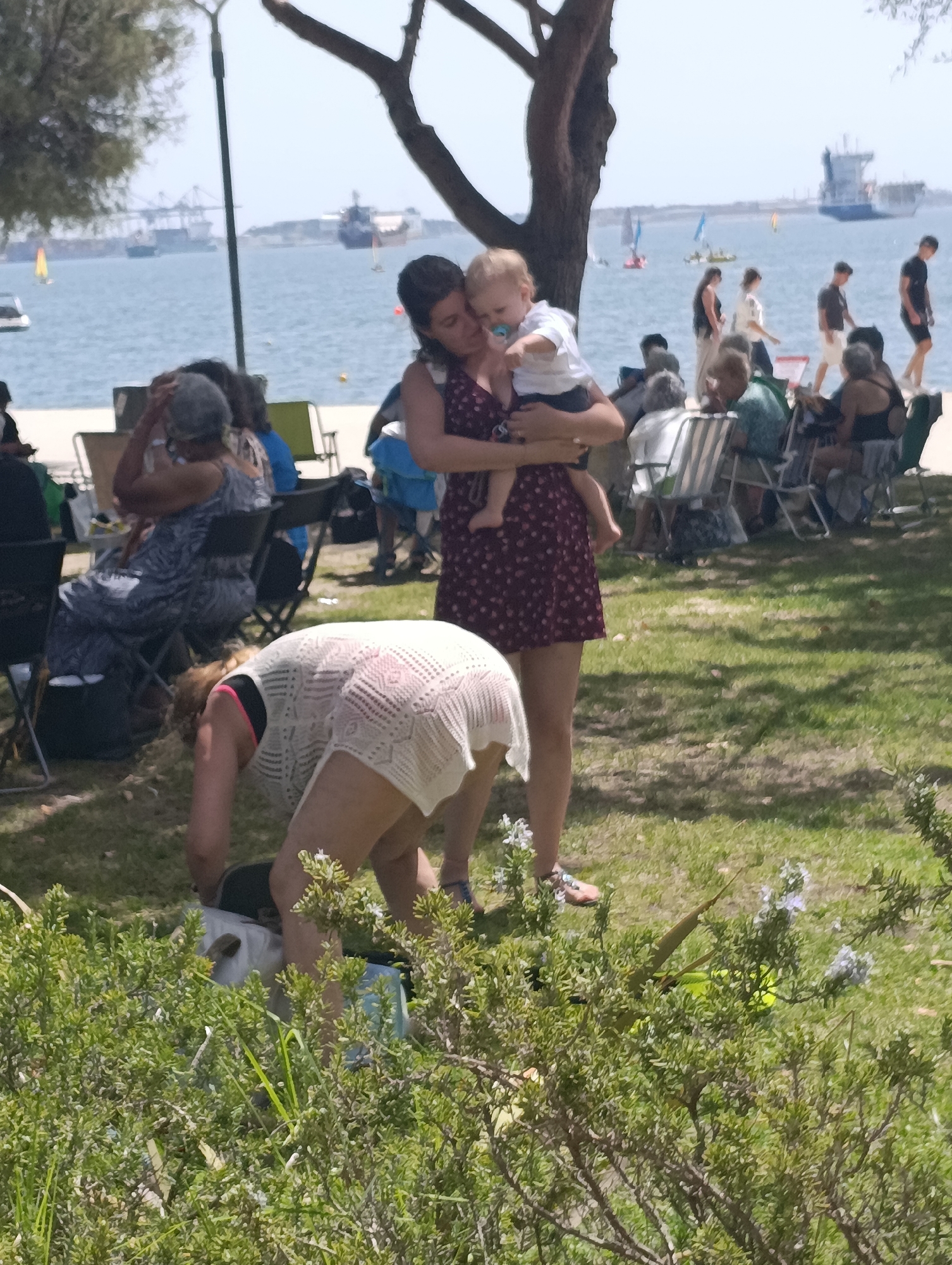 This is a medium-distance, horizontal-format, outdoor photograph captured on a sunny day. The scene depicts a group of people at a waterfront location, likely a park or promenade, with a body of water and boats in the background. In the foreground, a woman in a white lace cover-up is bending over, reaching into a light blue bag. Behind her, a woman wearing a dark floral print dress is holding a baby, who has a pacifier in its mouth. Several other individuals are seated on chairs and picnicking in the mid-ground. The environment is grassy with scattered trees and foliage. The background shows a harbor with several large ships and smaller sailboats on the water. The overall atmosphere suggests a casual gathering or outing.