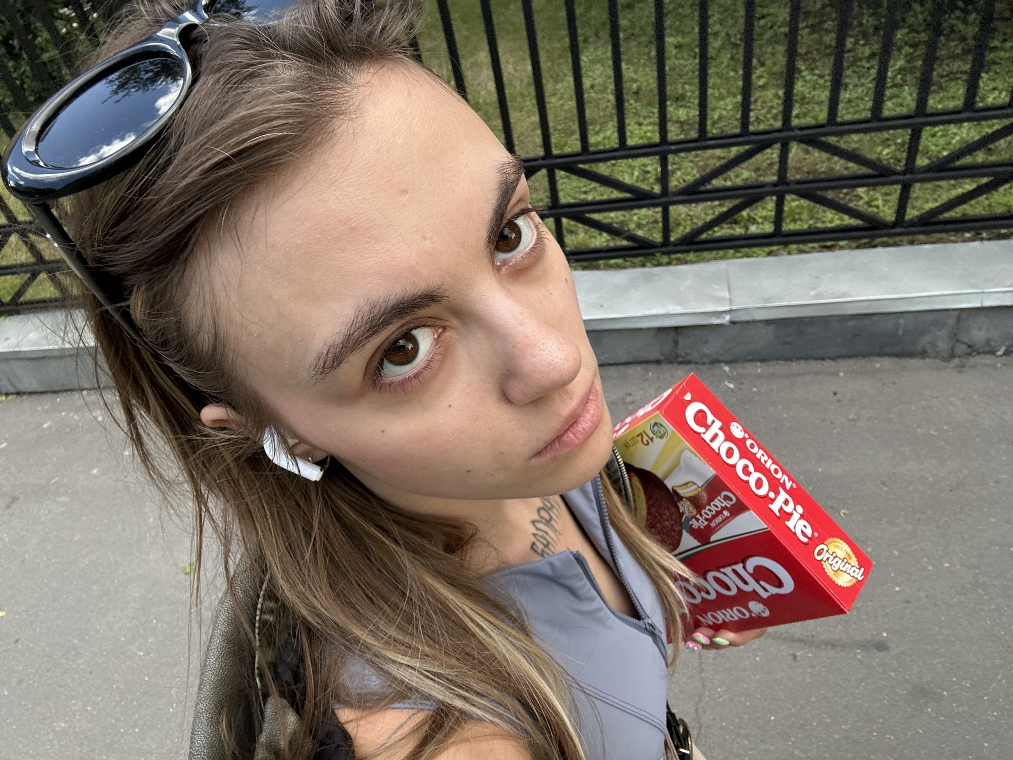 This is a first-person perspective selfie of a young woman outdoors, holding a red box of Orion Choco-Pie cookies. She has long, blonde-brown hair, is wearing sunglasses pushed up on her head, and has white earbuds in her ears. A tattoo is visible on her neck. She is wearing a grey zip-up top and a dark jacket. The background shows a black metal fence with a grassy area behind it, and a sidewalk or paved path in the foreground. The scene appears to be in a park or along a street in Moscow, Russia. The lighting suggests it is daytime, likely with overcast or diffused sunlight. The woman's expression is neutral. The text "ORION Choco-Pie Original" is clearly visible on the cookie box.