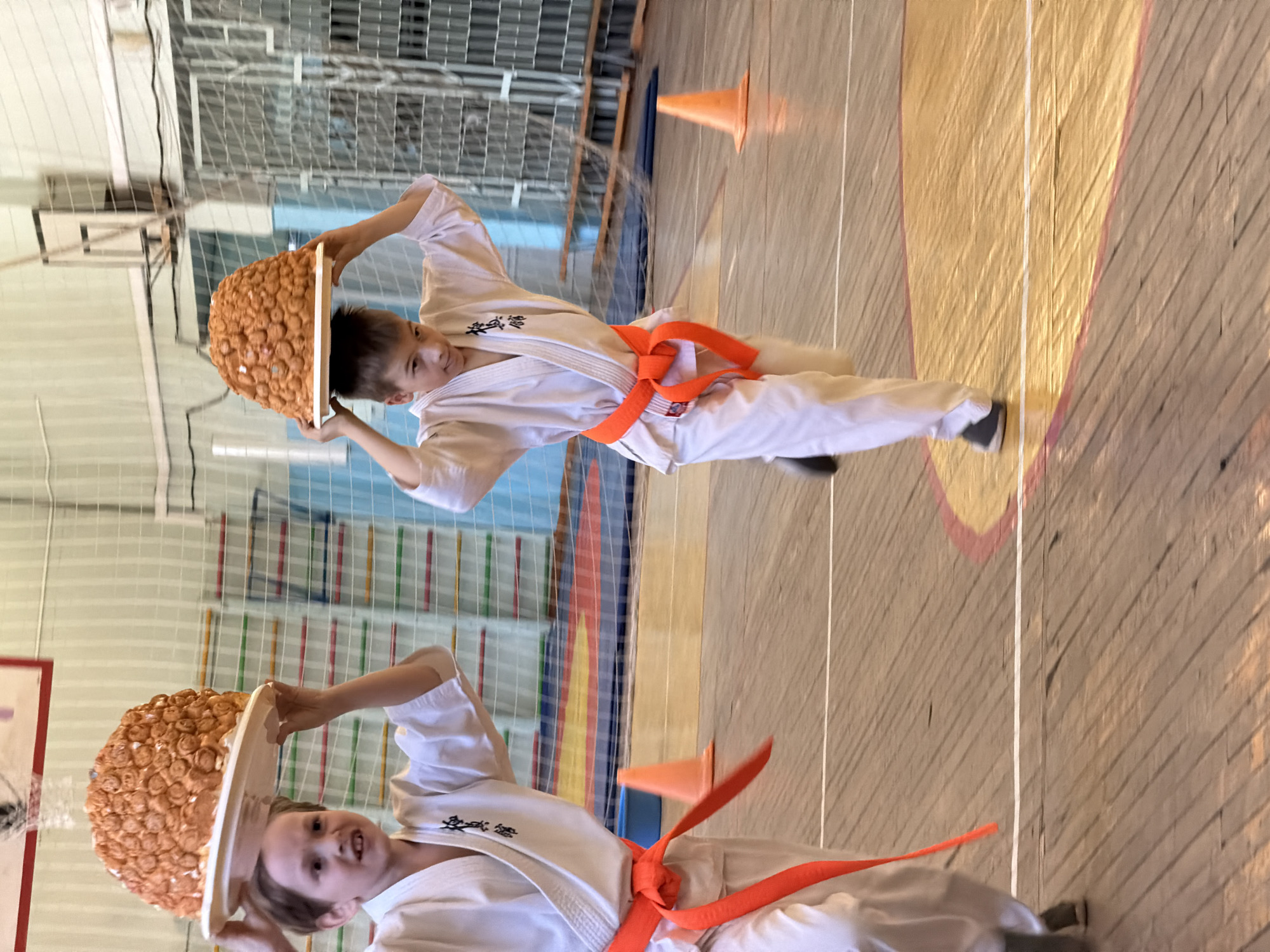 Two young boys in white karate uniforms with orange belts are participating in a game at what appears to be a karate event. They are each balancing a tray topped with numerous balls or round objects on their heads, seemingly navigating an obstacle course in a gymnasium. An orange cone is visible. The background shows typical gymnasium features like wall padding, a basketball hoop, and a metallic mesh structure. The floor is wooden and marked with lines. The location is Izhevsk, Russia, indicated by the setting and architecture.
