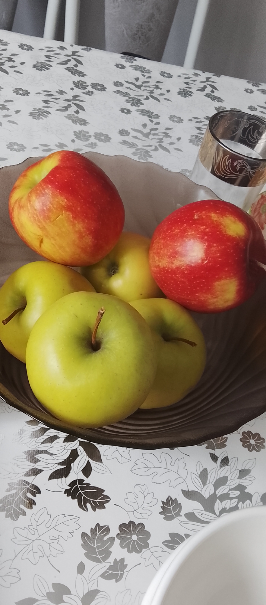 A bowl of apples sits on a table with a floral pattern. The bowl appears to be made of dark brown glass. Inside the bowl are five apples: three green apples and two red and yellow apples. One of the green apples is in the foreground, filling much of the frame. Behind it, two other green apples are visible, along with two red and yellow apples. To the right of the bowl, a glass with a decorative gold rim is partially visible. The table surface has a light-colored tablecloth with a dark gray floral pattern of leaves and flowers. The background is blurred, showing what appears to be furniture, possibly chairs, in shades of white and gray.