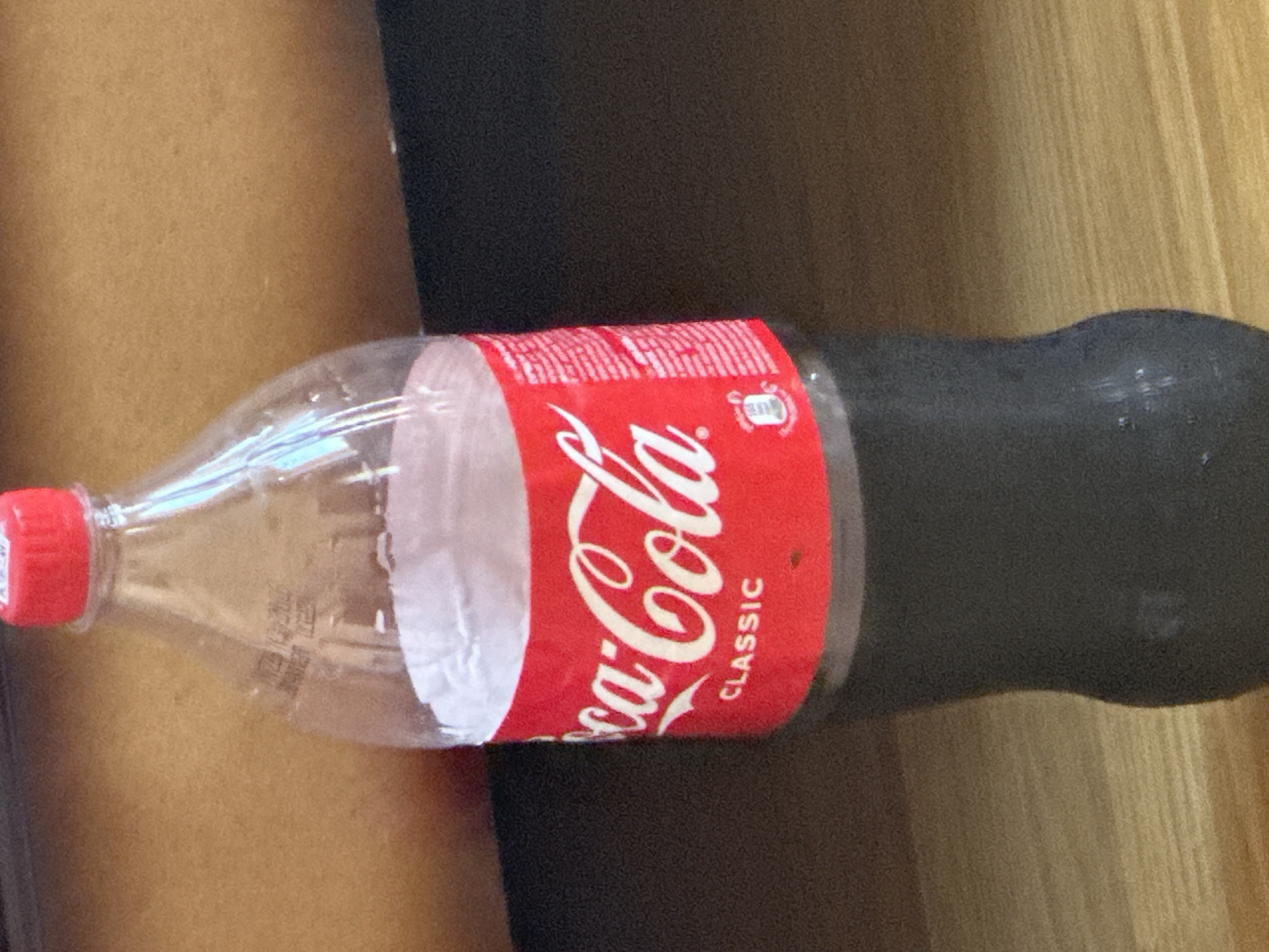 A partially filled plastic bottle of Coca-Cola Classic stands upright on a dark, reflective surface, likely a table. The bottle has a red cap and a white paper label with the iconic Coca-Cola logo in red and white script, along with the word "CLASSIC" below it. The liquid inside is dark brown, typical of cola. The background is a warm, light brown, appearing to be a wooden surface, possibly a wall or furniture. The lighting suggests an indoor environment, possibly a home or office. There are no people visible in the image, and no specific identifiable landmarks or signs that would indicate the city of Gyumri, Armenia. The image is a close-up of the bottle, with the focus primarily on the product itself.