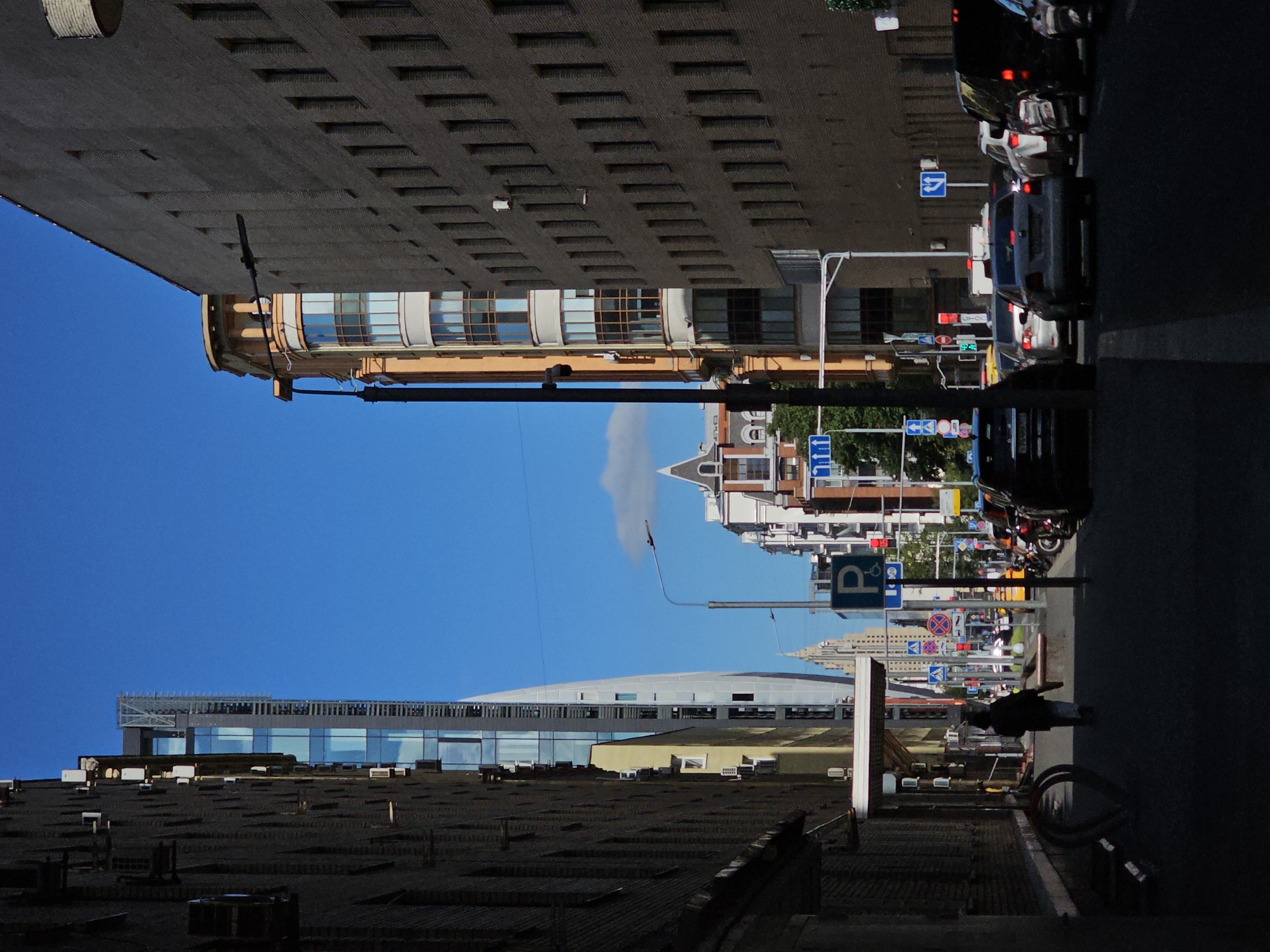 The image depicts a street scene in Moscow, Russia, under a clear blue sky with a few scattered clouds. Tall buildings, some with modern glass facades and others with older brickwork, line both sides of the street, creating a canyon effect. A lone figure walks away from the viewer down a shadowed sidewalk. Several cars are visible on the street, some in traffic, suggesting a typical urban environment. Various street signs, including a parking sign with a wheelchair icon, are present. The overall impression is one of a busy city street during daylight hours.
