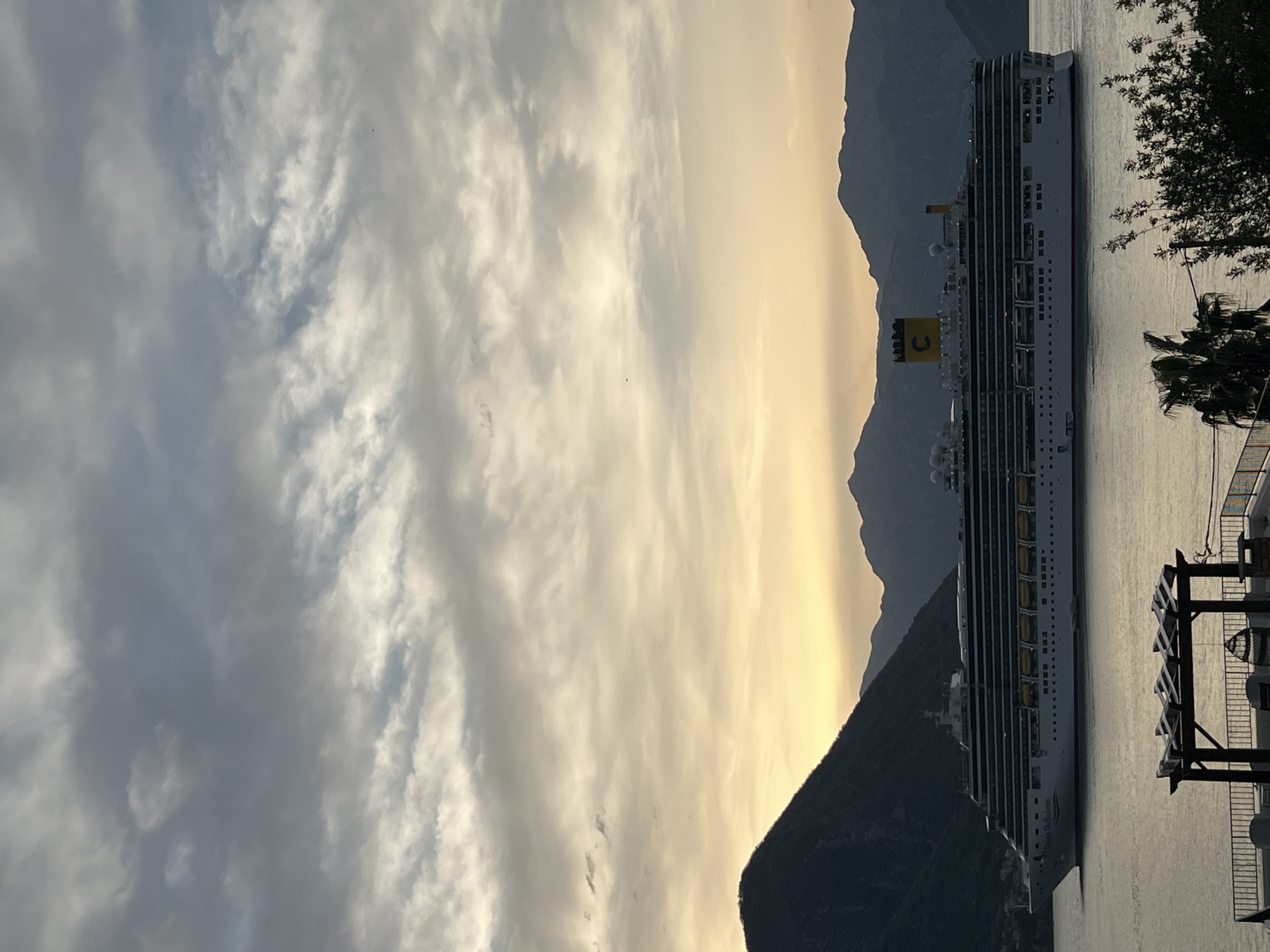 The image captures a Costa cruise ship docked in the Bay of Kotor, off the coast of Dobrota, Montenegro. The ship is positioned vertically on the right side of the frame, revealing its multiple decks and the yellow "C" logo on its funnel. Mountain silhouettes rise behind the ship, with a sky filled with clouds transitioning from dark gray to a golden hue toward the mountains. The water is calm, reflecting the sky's colors. On the right, a pier with wooden structures and palm trees is visible.