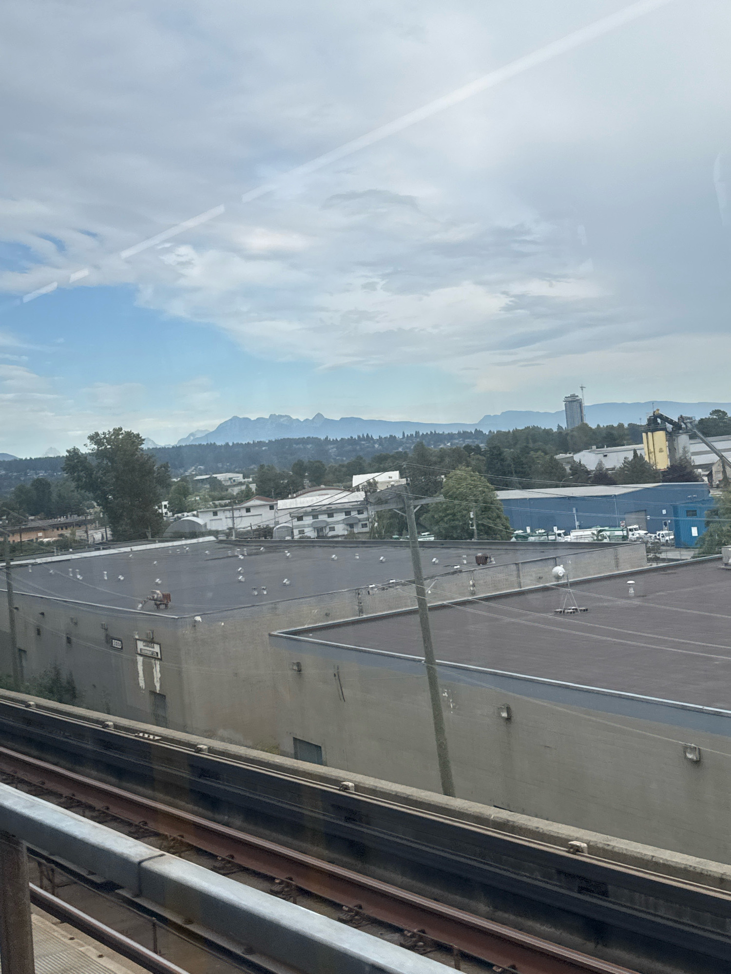 This is a medium-distance, eye-level, outdoor, horizontal photograph taken from a moving train in New Westminster, Canada. The scene depicts an urban industrial area with a mountain range visible in the background under a cloudy sky. In the foreground, train tracks and a concrete barrier are visible, suggesting the viewpoint is from a SkyTrain.
Dominating the mid-ground are several flat-roofed industrial buildings. The closest building, on the left, is a light-colored structure with a darker, grey flat roof. Visible on its facade are two rectangular, white signs with black lettering, one partially obscured, and what appear to be ventilation units or skylights. Another larger, grey building with a dark, flat roof is situated behind it, also featuring what appear to be HVAC units or vents. Further into the mid-ground, a long, pale blue industrial building is present, with a few white trucks parked outside. Adjacent to it is a yellow building with a crane extending from its top.
The background features a dense line of trees separating the industrial area from a distant mountain range, which is partially obscured by haze. The sky is overcast with scattered clouds, with a hint of blue peeking through. The overall impression is that of an industrial landscape on a hazy day.