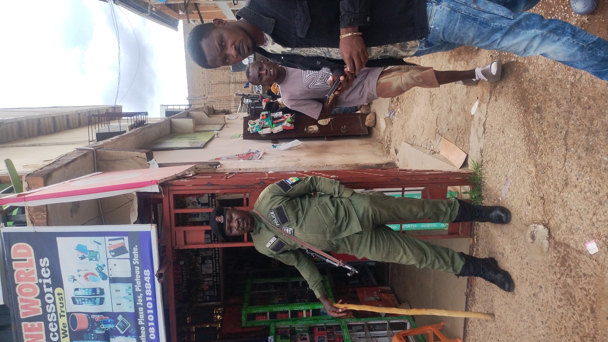 The image depicts a street scene in Jos, Plateau State, Nigeria, specifically in the vicinity of Mintheo Plaza and Ahmadu Bello Way, as indicated by visible signage. The scene is captured at eye level on an overcast day.
In the foreground, a male individual of dark complexion stands to the right, facing towards the camera. He wears a black denim jacket over a patterned t-shirt, blue jeans, and light-colored shoes. A gold-colored chain bracelet is visible on his left wrist. He holds two black smartphones in his hands, with his gaze directed forward.
To the left, another male individual, identified by a name tag as "CHISOM MBONU," stands partially inside a shop doorway. He is a dark-complexioned adult wearing a dark green long-sleeved uniform, black combat boots, and a black beret. His uniform features a "POLICE" patch on the left chest and a Nigeria Police Force (NPF) logo on the right shoulder. An AK-style assault rifle, black in color, is slung across his chest with a black strap. He holds a light-colored wooden staff vertically in his left hand, resting it on the ground, while his right hand is in his pants pocket.
Behind the foreground figures, a third male individual, also of dark complexion, stands in a narrow dirt and concrete alleyway. He wears a light grey short-sleeved t-shirt with a dark graphic and patterned brown shorts, holding a dark rectangular object in his right hand.
The shop behind the uniformed officer features a partially open red metal grate door. Inside, shelves display various electronic items, likely phones and accessories. Above this shop, a large blue banner advertises "ONE WORLD Accessories," displaying images of various phone-related products, with an address of "Mintheo Plaza Jos, Plateau State" and a contact number. Below it, a smaller sign provides an address on "Ahmadu Bello Way" and another phone number.
The ground consists of an uneven mix of concrete and packed dirt, with some scattered debris, including a white plastic wrapper, near the officer's feet. The surrounding buildings are multi-story, constructed of light-colored, possibly painted concrete, showing signs of wear and age. Some buildings feature visible balconies with metal railings. The sky above is uniformly grey and overcast.
