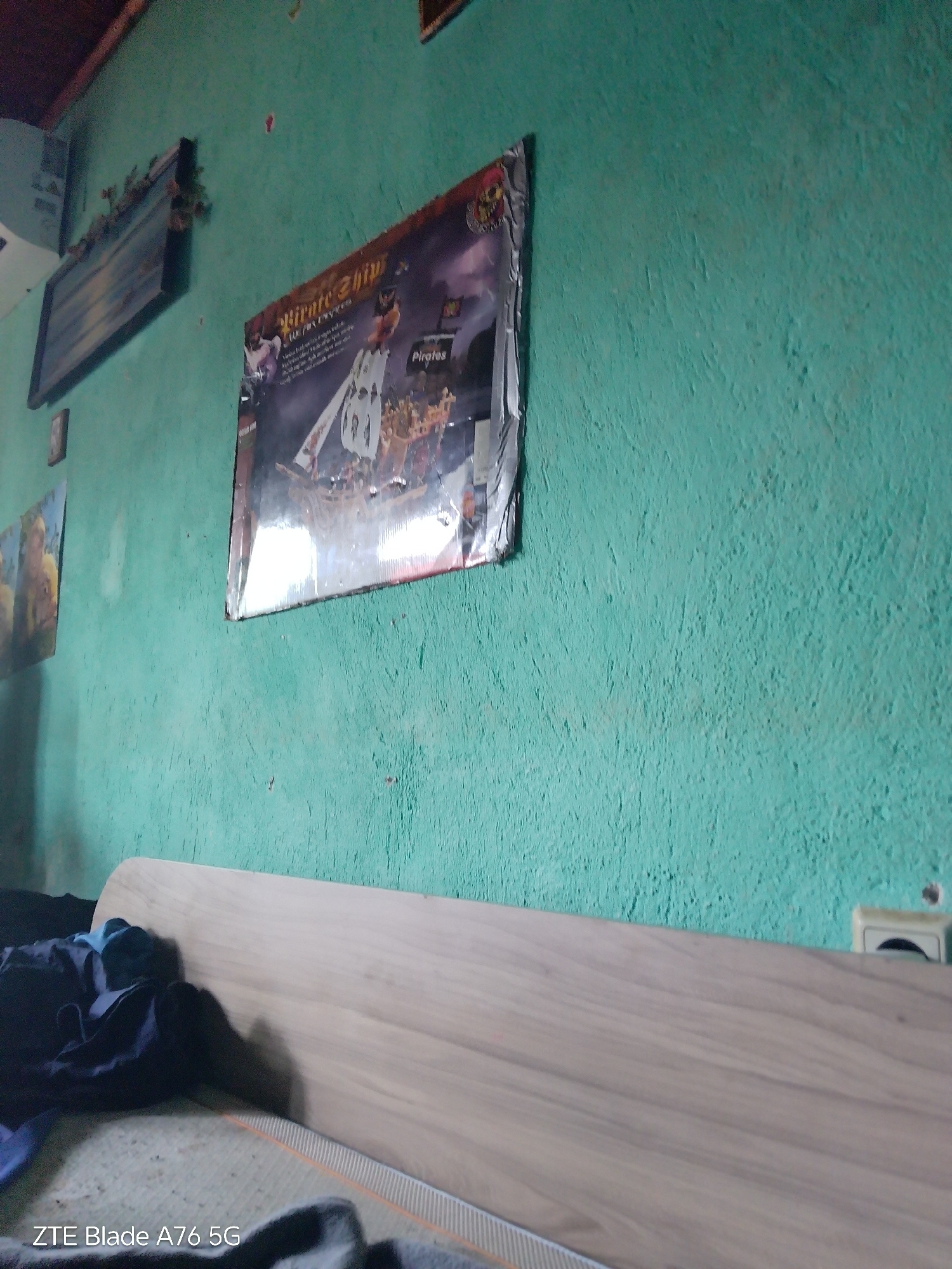 The image shows an indoor scene with a rough, turquoise-painted wall. On the wall, a large poster featuring a pirate ship and cartoon-like figures is displayed. To the left of the pirate poster, a smaller framed picture is hung, and further to the left, a rectangular object with some greenery is mounted on the wall. A light-colored wooden headboard is visible in the foreground, partially covered by dark clothing. The floor appears to be a mat with a striped pattern. The bottom left corner of the image displays text that reads "ZTE Blade A76 5G". The scene is likely an interior of a room. There is no visible information in the image to indicate its specific location within Stara Zagora, Bulgaria.