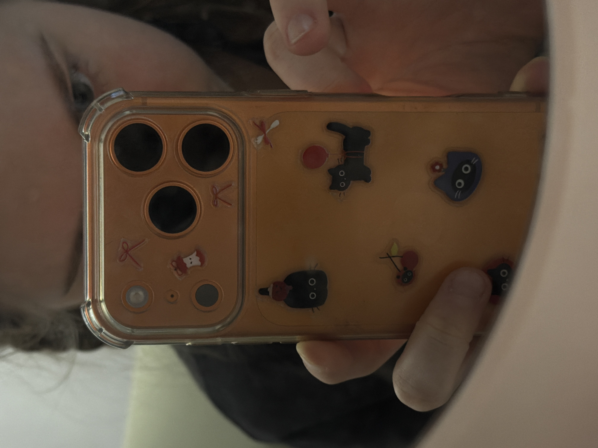 A person holds an orange cell phone case with cartoon cat stickers on it. The phone is positioned in front of the person's face, partially obscuring it. The background is a blurry mix of white and dark colors. The image does not provide any information to determine the location in Perm, Russia.