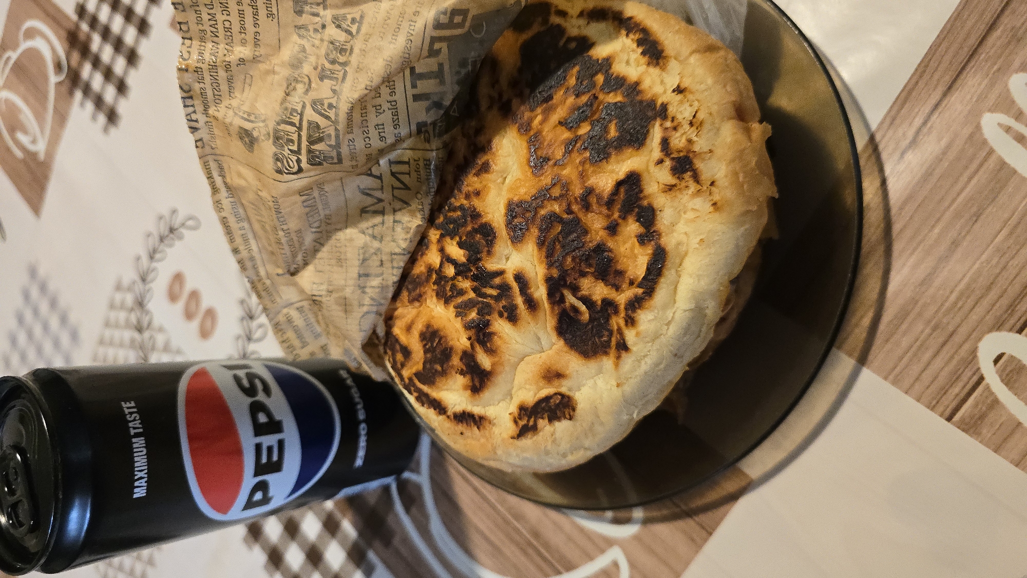 A black Pepsi can with "MAXIMUM TASTE" and "ZERO SUGAR" printed on it is placed on a table with a checkered and floral pattern tablecloth. Next to the can is a paper-wrapped sandwich, partially unwrapped, revealing a toasted bread bun with dark char marks. The sandwich is on a dark glass plate. The image appears to be taken indoors.