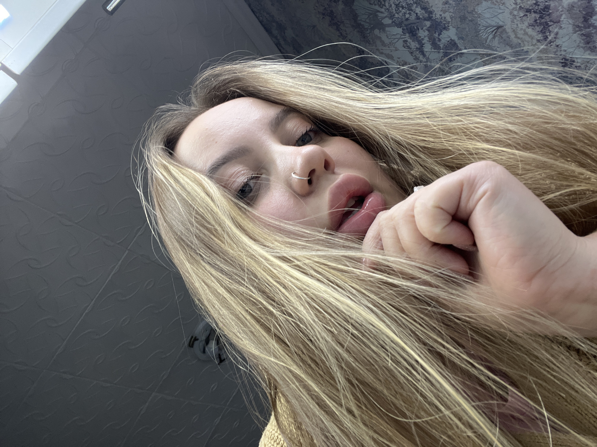The image captures a close-up, low-angle perspective of a young woman with long, flowing blonde hair, looking directly into the camera. Her face fills a significant portion of the frame, emphasizing her features. She has light-colored eyes and a small silver hoop nose ring in her left nostril. Her lips are full and slightly parted, with a glossy appearance, and her tongue is subtly visible. One of her hands is partially visible on the right side of the frame, near her mouth, with fingers lightly curled or touching her hair.
The setting appears to be indoors. The background consists of two distinct sections: the upper left shows a plain, textured gray surface, possibly a ceiling or wall, with a rectangular light fixture visible at the very top edge. The upper right and behind her head features a darker, patterned wallpaper or ceiling design with what appears to be a subtle floral or leafy motif. The low-angle shot creates a dynamic and engaging portrait, suggesting an intimate or direct interaction with the viewer. No text is visible in the image. The location context provided is Novopokrovskaya, Russia, though no specific visual cues within the image confirm this.