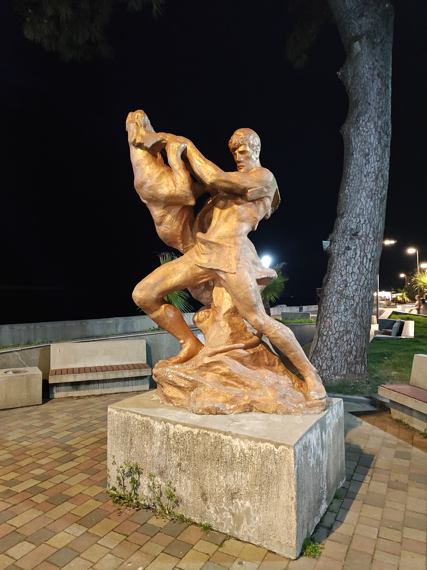 Captured at night in an urban setting in the city of Unknown, Unknown, a bronze-colored statue commands attention. The statue depicts a man carrying or lifting another figure, possibly in distress or rescue. The artwork rests upon a square concrete base, which is positioned on a patterned brick walkway. Near the statue, there are benches situated alongside a concrete retaining wall and a tree. In the background, faint lights suggest a waterfront or promenade area.