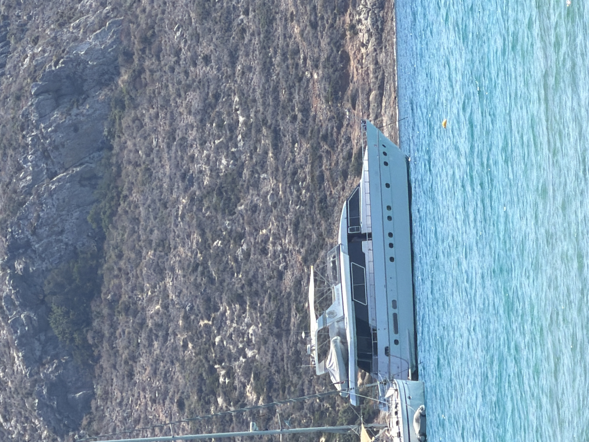 A white motor yacht is docked in the turquoise waters of Greece, with a rocky, scrub-covered hillside in the background. The yacht is a sleek, modern vessel with multiple windows, a covered deck, and a smaller dinghy secured to its side. A sailboat mast is partially visible on the left, indicating other vessels are present in the bay. The water is clear and a bright azure, with gentle ripples on the surface. The hillside is steep and rugged, covered in low-lying dry vegetation and scattered rocks. The sky is a pale blue, suggesting a sunny day. The scene depicts a tranquil coastal setting in Greece.