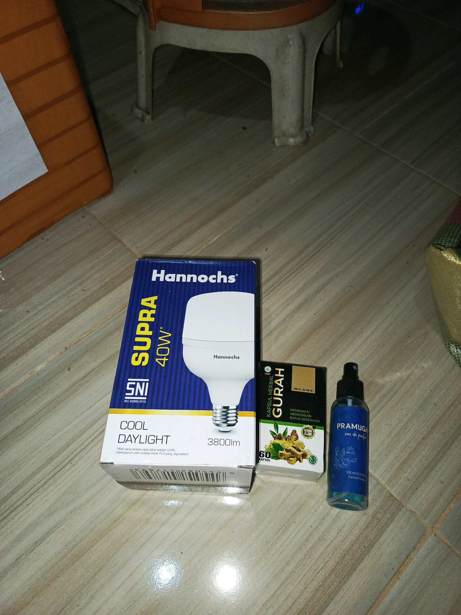 In Megang Sakti I, Indonesia, an image captures a collection of items on a tiled floor, illuminated by artificial light. In the foreground, a blue and white cardboard box for a "Hannochs SUPRA 40W" light bulb is partially visible. To the right of the light bulb box are two smaller items: a dark box labeled "KAPSUL HERBAL GURAH" featuring an image of ginger, and a blue spray bottle of "PRAMUGAR eau de parfum". A portion of an orange object, possibly a box or cushion, is visible in the upper left corner, and the legs of a light-colored stool or table are in the upper center. The scene is set indoors, with the light reflecting off the glossy floor tiles. No individuals are present in the frame.