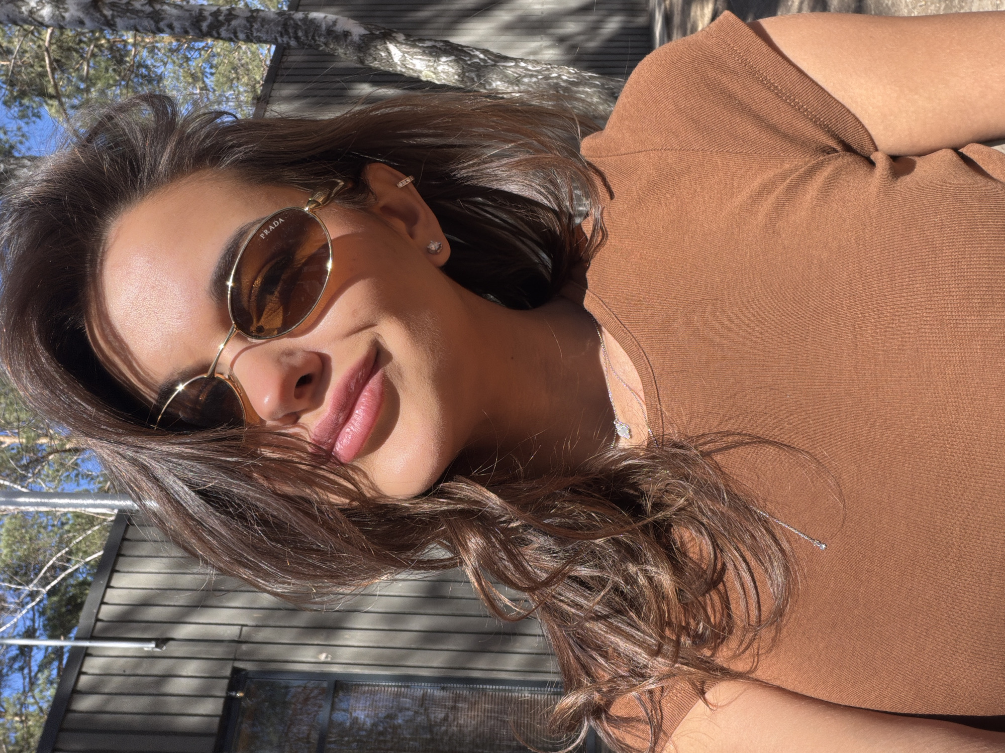 A young woman is depicted in a close-up, rotated portrait under bright daylight in what appears to be an outdoor setting in Krasnoyarsk, Russia. She has long, wavy dark brown hair that cascades around her face and shoulders. Her lips are slightly parted in a gentle smile, revealing a natural pink hue. She wears large, oval-shaped sunglasses with gold frames, and the brand name "PRADA" is clearly visible on the upper left lens.
Her attire consists of a simple, fitted, short-sleeved top in a warm, earthy brown color, possibly ribbed. Accessories include a small, sparkling stud earring on her earlobe and a thin cuff earring higher up on her ear cartilage. A delicate silver chain necklace with a small pendant can be seen resting on her chest.
The background shows a clear blue sky and the branches of pine trees. Partially visible behind her is a modern-style building with dark, horizontal wooden or paneled siding and a reflective window. The strong sunlight casts distinct shadows, indicating a bright, clear day. The overall mood conveyed by her smile is one of contentment and relaxation, possibly enjoying the sunny weather outdoors.