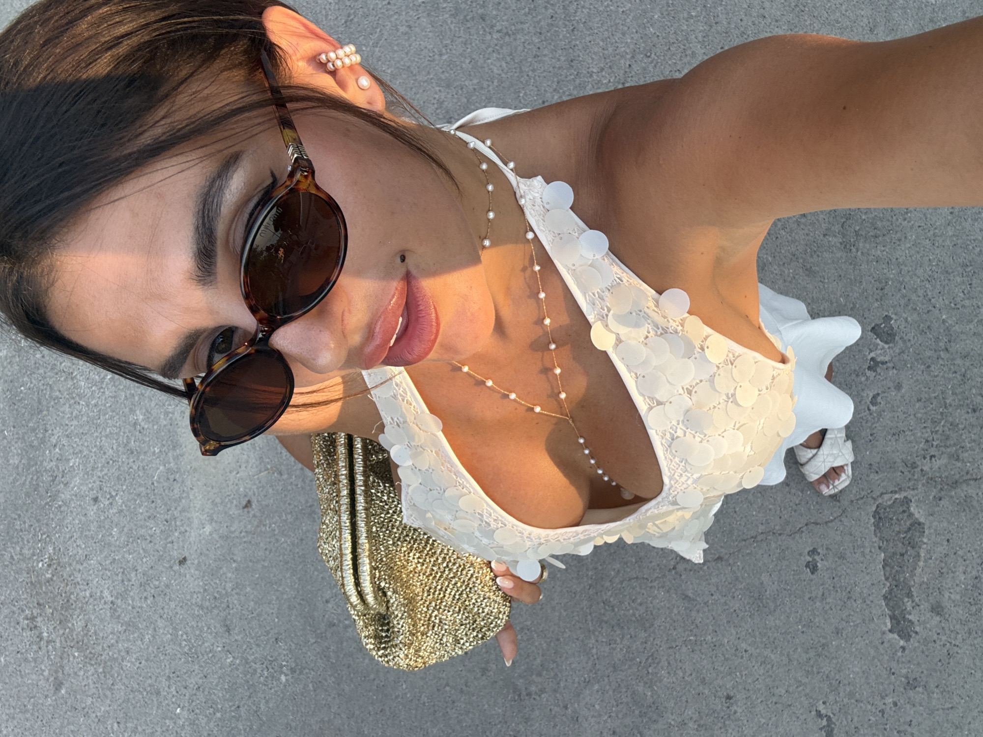 The media file shows a first-person perspective of a person holding a smartphone and taking a selfie. The subject is a woman with dark hair, wearing sunglasses and a white sequined top. She is accessorized with pearl earrings and a delicate pearl necklace. She is also holding a sparkly gold clutch bag. In the background, the ground appears to be concrete, with some cracks and marks. The lighting suggests it is daytime, with sunlight casting shadows. The woman's makeup is visible, with defined eyebrows and lipstick. She is wearing white sandals. The scene is likely outdoors, given the natural lighting and the textured ground. The overall impression is one of style and leisure, possibly during a vacation or a social outing. The location context of Fethiye, Turkey, suggests a warm-weather destination.