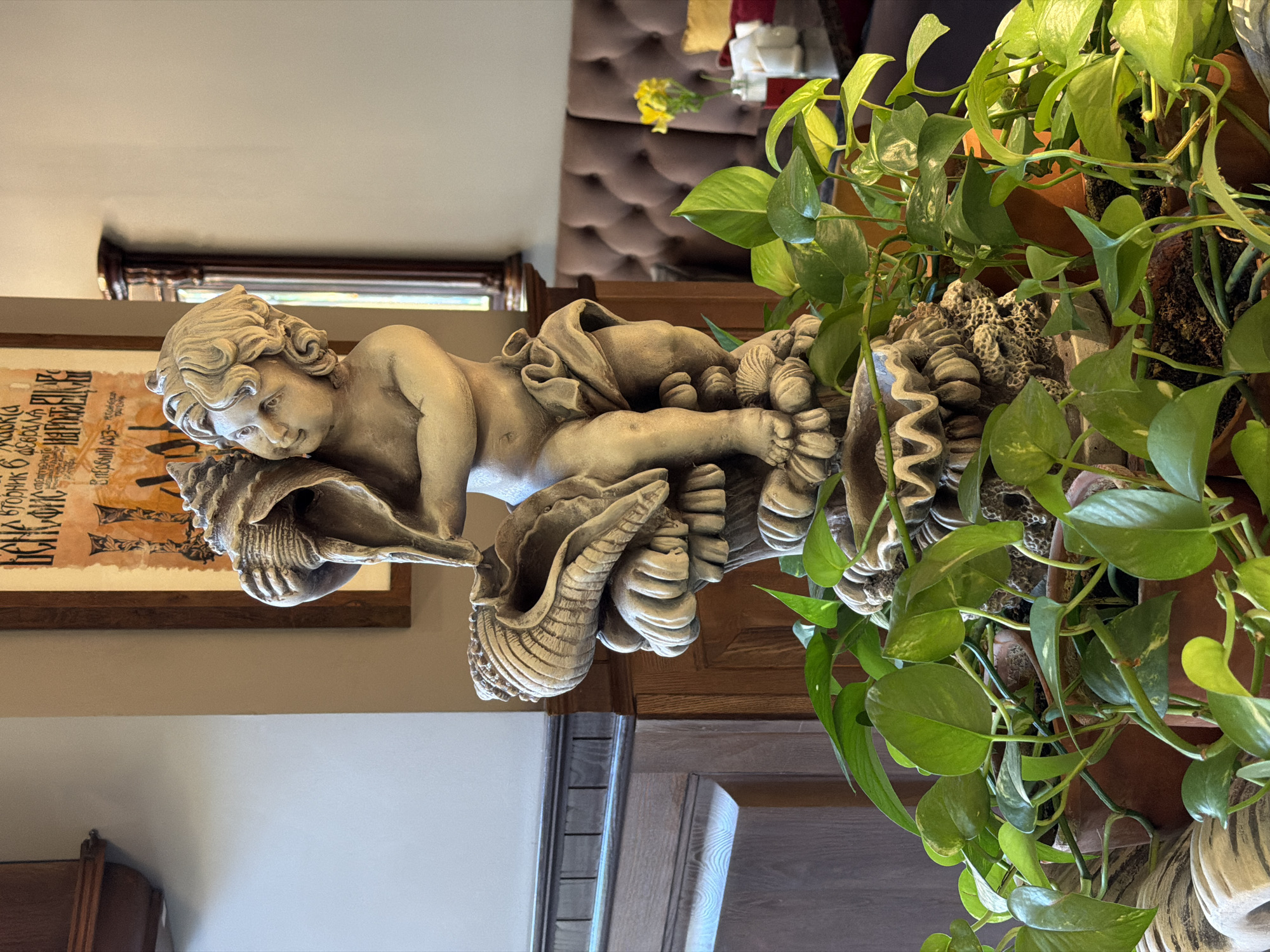 The image depicts a stone cherub fountain in the foreground, adorned with seashells, surrounded by lush green plants. In the background, a tufted brown sofa is visible, with a yellow flower in a vase on a table. A framed artwork hangs on the wall behind the fountain. The scene appears to be an indoor setting, possibly a cafe or restaurant. The location is not identifiable from the image alone and cannot be determined to be Rostov-on-Don, Russia.
