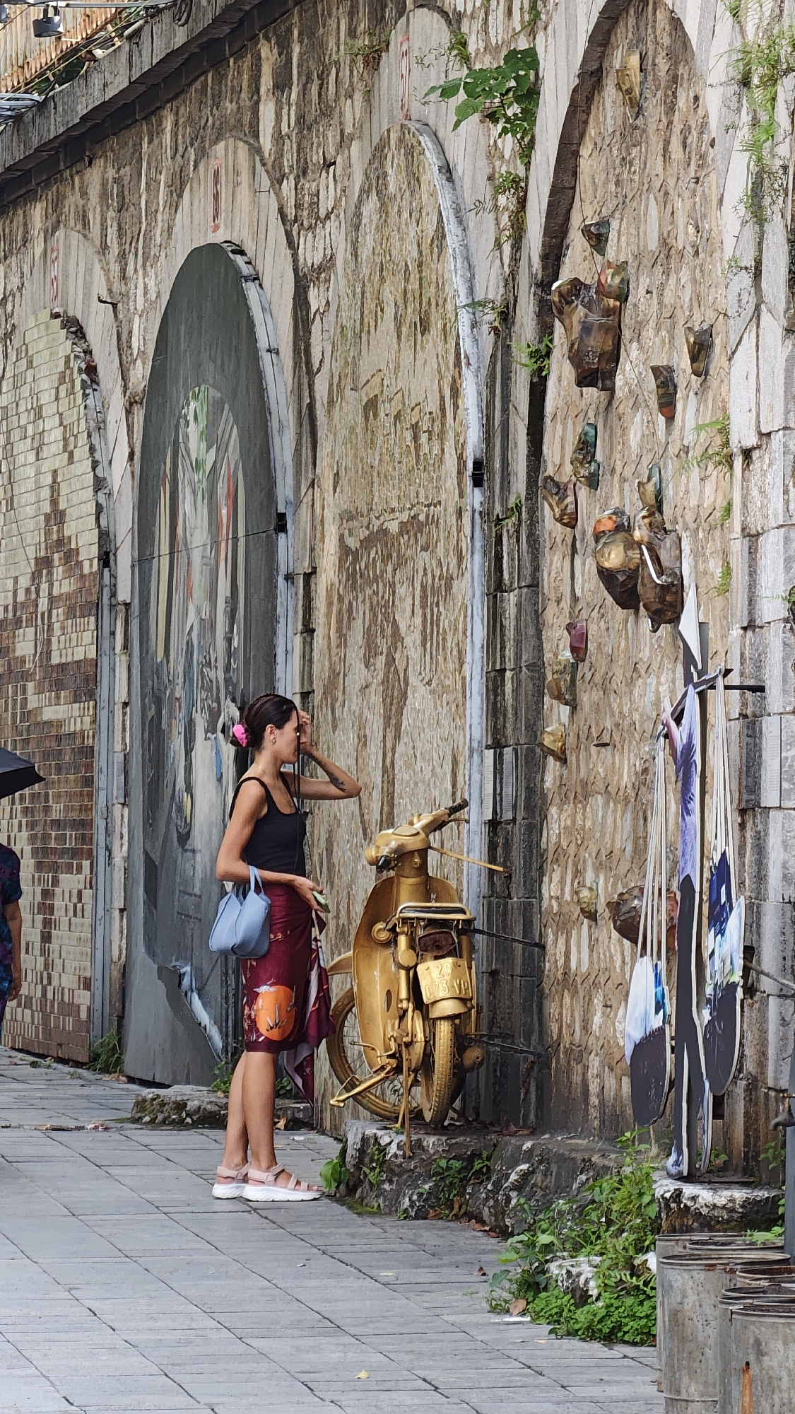 The image shows a young woman standing on a tiled sidewalk next to a textured stone wall with arched alcoves, likely in Hà Nội, Vietnam. The wall features artwork, including a faded mural and mounted metal sculptures. The woman is wearing a black tank top, a patterned wrap skirt, and pink sandals. She has a light blue purse and is touching her forehead with one hand. A gold-painted vintage motorcycle is parked near her, and a rack displaying items that appear to be photographs is visible to the right of the motorcycle. The environment includes foliage growing near the base of the wall and metal containers in the foreground. A blurred figure with an umbrella is partially visible to the left, indicating the presence of other pedestrians.