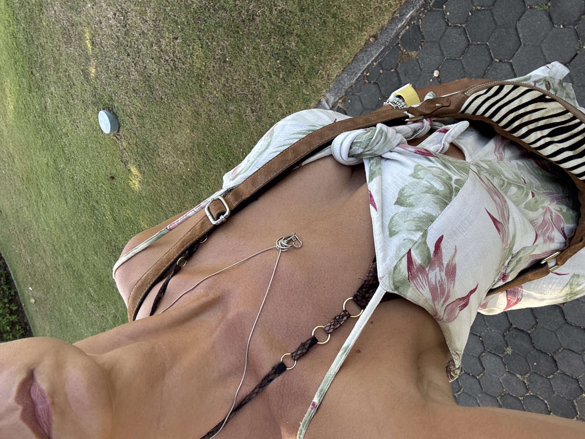 A woman stands on a paved path near a grassy area. She is wearing a floral print dress and a brown shoulder bag with a zebra print accent. A silver necklace with a heart-shaped pendant is visible around her neck. In the background, a light blue cap or lid rests on the grass. The scene appears to be in Kuta Selatan, Indonesia, based on the provided context.