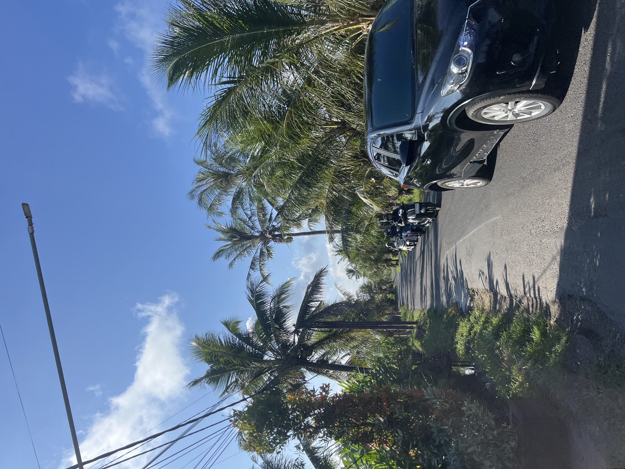 The image depicts a street in Sukawati, Indonesia. A black car is parked on the right side of the asphalt road. A row of scooters is parked behind the car. Palm trees line the side of the road. The sky is blue with scattered white clouds. Utility poles and power lines are visible on the left side of the frame.