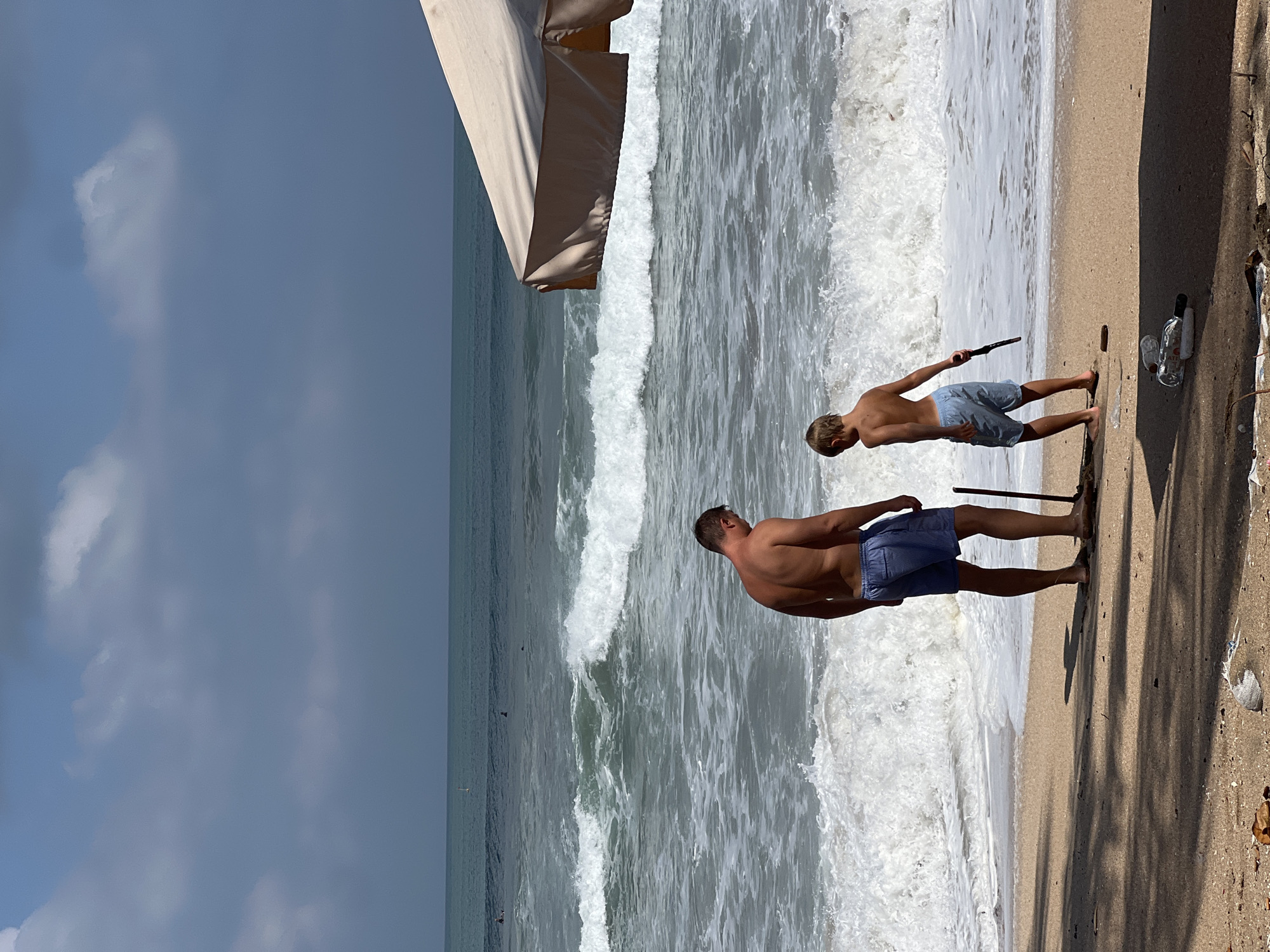 In Kuta, Indonesia, two shirtless individuals, an adult male and a young boy, stand on a sandy beach facing the ocean. The man, wearing blue shorts, holds a dark-colored pole. The boy, also in blue shorts, holds a black object in his right hand. They are positioned between the wet sand and the dry sand, with the ocean waves crashing behind them. The water is a deep blue-green, and white foam indicates active surf. In the background, a large, light-colored beach umbrella provides shade. The sky is partly cloudy, with patches of blue visible. The foreground of the beach contains scattered debris, including plastic bottles and leaves, casting shadows from unseen trees or structures.