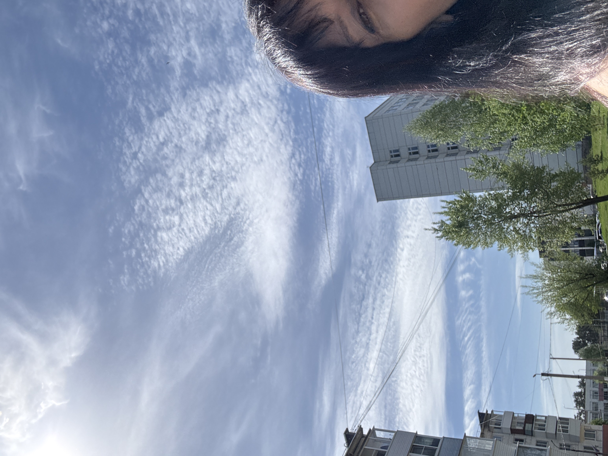 The image shows a selfie taken outdoors in Chaikovsky, Russia, partially capturing a person's forehead, eyes, and dark hair in the upper part of the frame. The subject is looking up at the sky, which is filled with wispy, cirrus-like clouds against a blue background. A high-rise, light-colored building and trees with green foliage are visible in the middle right. Power lines run through the scene. Lower in the frame are other multi-story buildings. The brightness suggests it is daytime.
