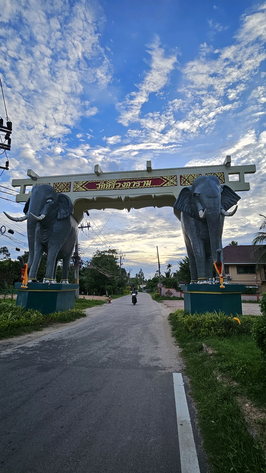 The image depicts an archway adorned with decorative Thai script, flanked by two monumental elephant statues. The archway spans over a paved road, with a lone motorcyclist visible in the distance, riding away from the camera. Lush green vegetation and trees line the sides of the road, and a building with a tiled roof is situated to the right. The sky above is a vibrant blue, filled with scattered white clouds, suggesting it may be late afternoon or early evening. The overall scene suggests an entrance to a significant location, possibly a temple or a landmark in Ko Samui District, Thailand.
