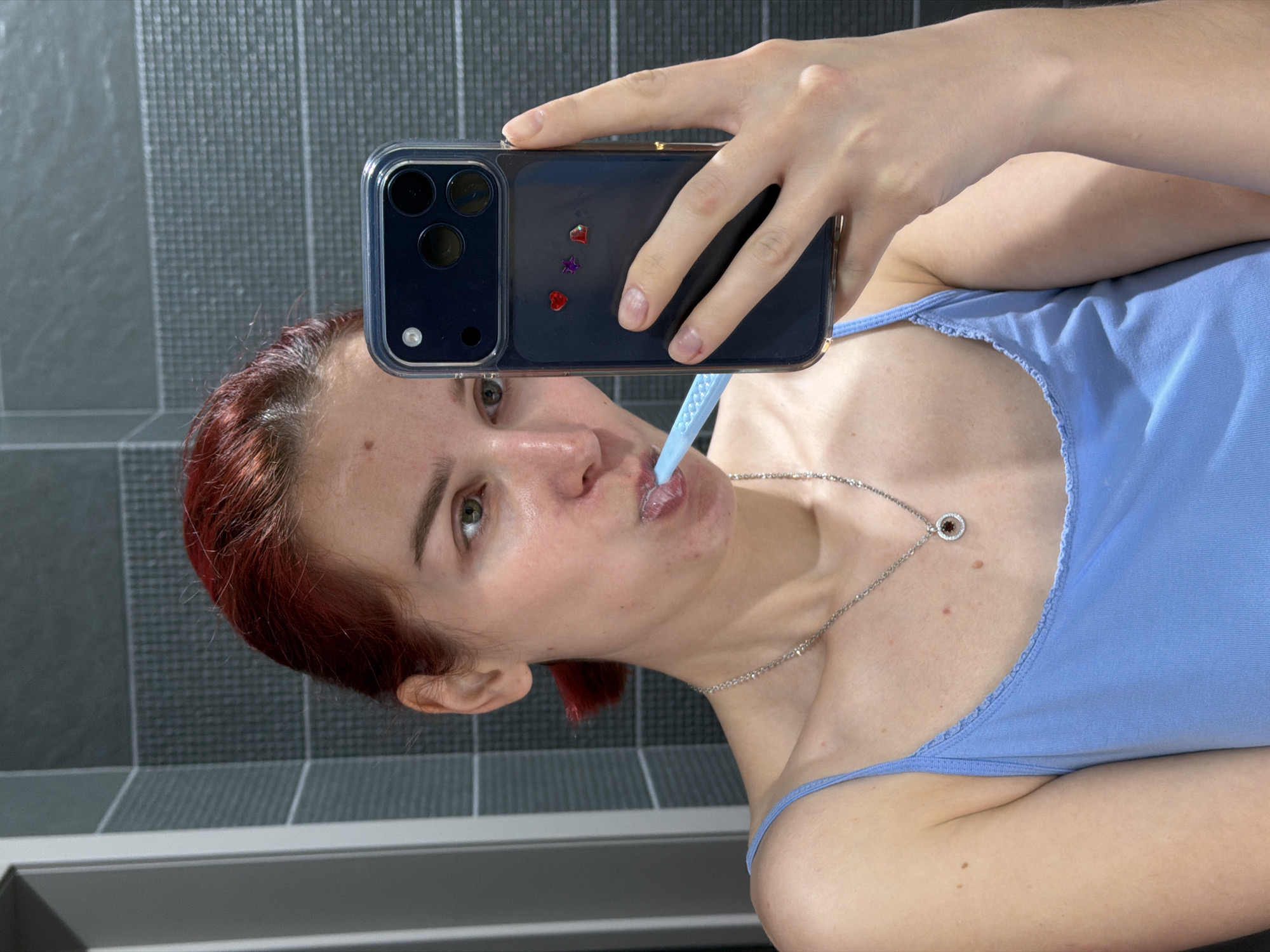 A young, light-skinned woman with red hair is taking a selfie in what appears to be a bathroom. She is holding a blue smartphone with a clear case adorned with small, colorful heart and star stickers. The phone is positioned so that the camera is facing her and capturing the shot. She is brushing her teeth with a light blue toothbrush. She is wearing a light blue tank top and a silver necklace with a circular pendant.
The background features a tiled wall with alternating patterns of dark gray and light gray rectangular tiles. A strip of light gray trim runs along the bottom of the tiled wall.
There is no information to indicate the location is in Chaikovsky, Russia.