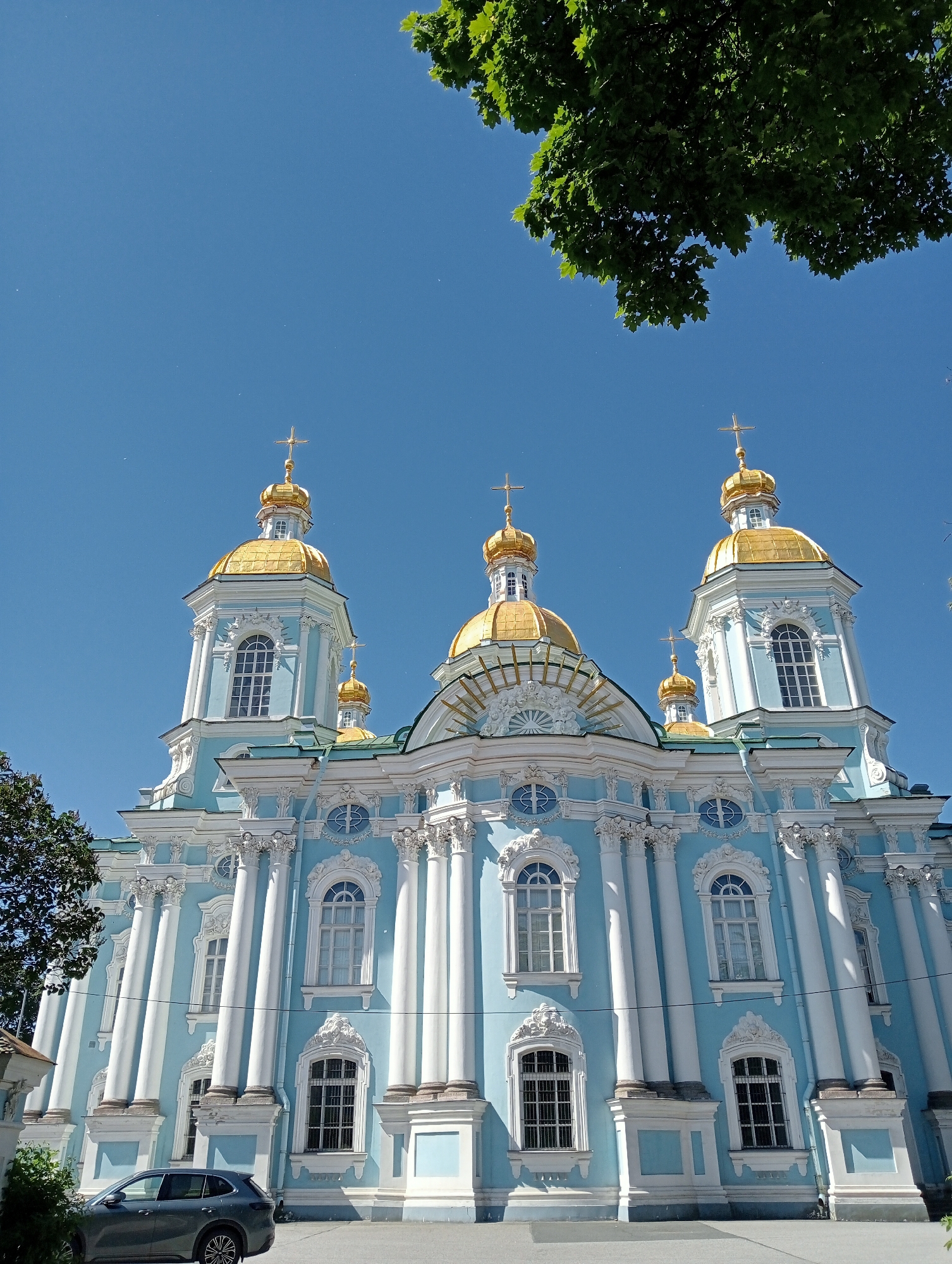 This media file shows a low-angle shot of the Saint Nicholas Naval Cathedral in Saint Petersburg, Russia. The sky is a clear, vibrant blue, suggesting a bright, sunny day. Lush green leaves from a tree are visible in the upper right corner of the frame, adding a natural element to the scene.
The cathedral itself is a striking architectural masterpiece, painted a vivid sky blue and adorned with white ornate details and fluted columns. Its prominent feature is the series of gilded domes, each topped with a cross, which catch the sunlight. The intricate stonework around the windows and cornices showcases classical design elements.
In the lower left of the image, a modern, dark grey SUV is parked on a paved surface, providing a contemporary contrast to the historic cathedral. Some greenery from bushes or small trees is visible on the far left, partially obscuring a small portion of a white building.
The low angle emphasizes the grandeur and height of the cathedral, making it appear monumental against the expansive blue sky. The image captures a moment of stillness, a snapshot of a beautiful day in a historic city. No people are visible, and there is no visible text within the frame.