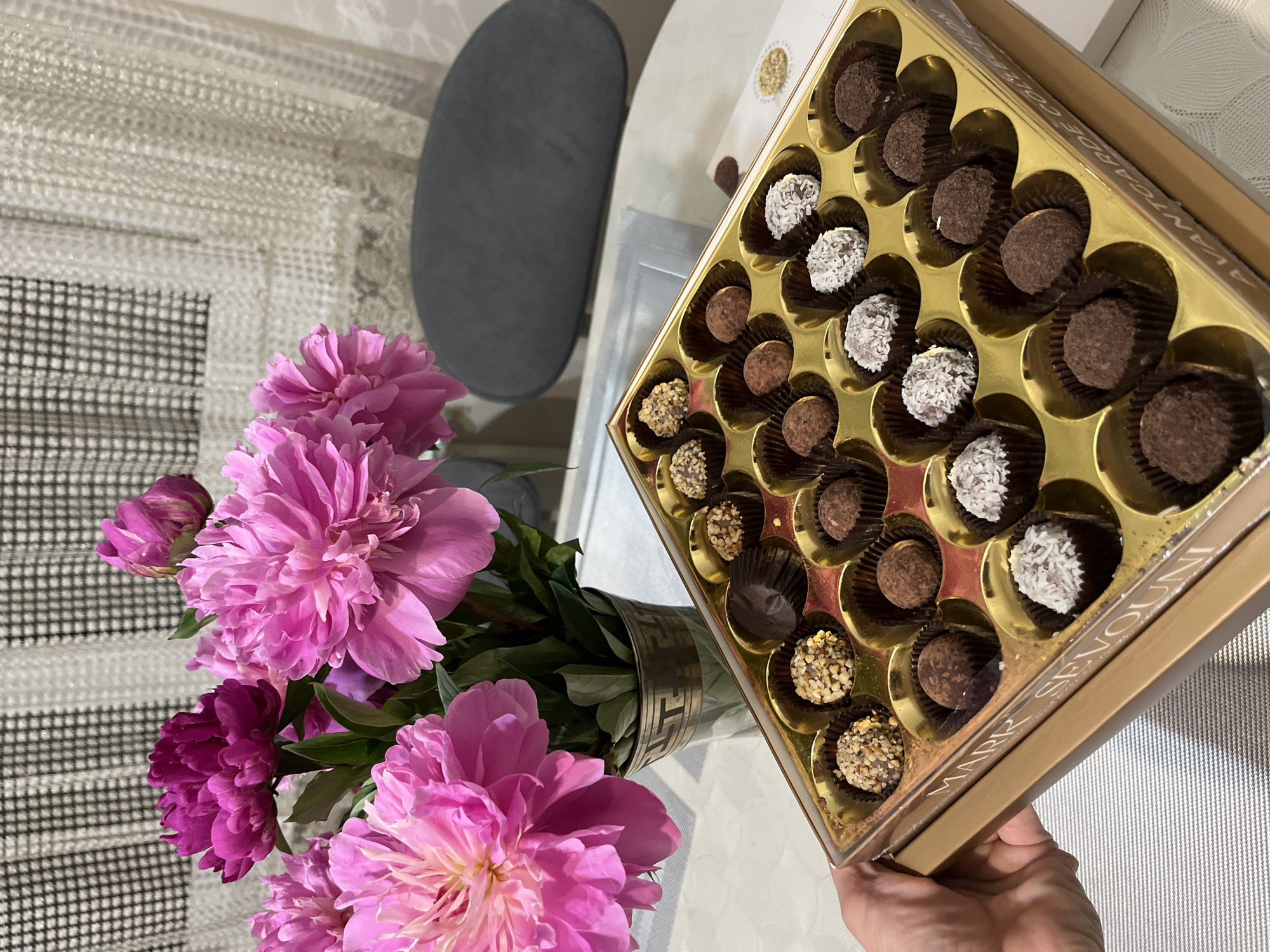 A high-angle, eye-level shot shows a hand holding a gold box of chocolates beside a vase of pink peonies. The chocolates are of various types, some coated in coconut flakes, some in chopped nuts, and some are plain chocolate. The box appears to be a gift box. The flowers are in a metal vase with a decorative band. The scene is set on a table. The location cannot be determined based on the image. The city of Kaspiysk, Russia, is not identifiable.