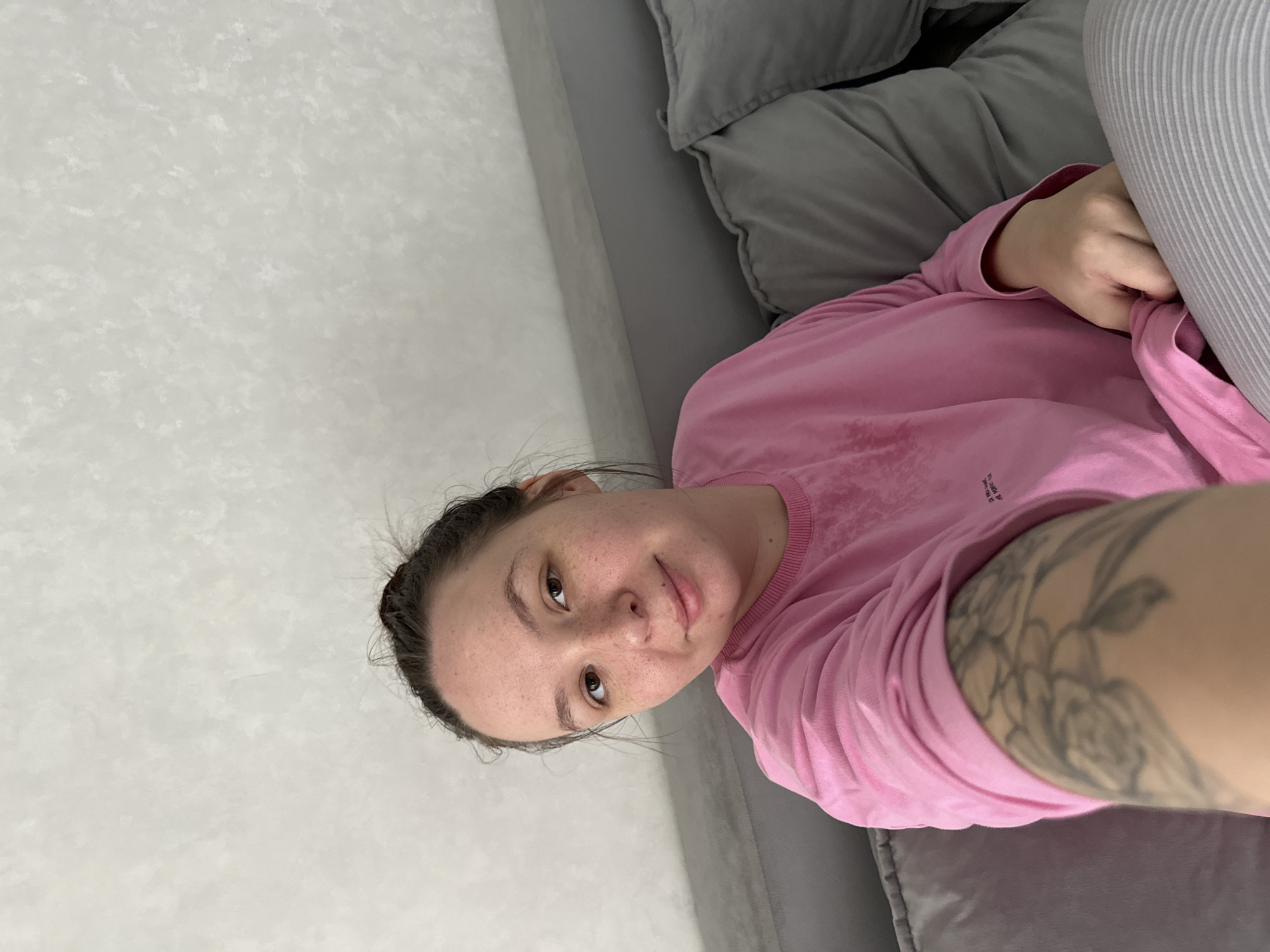 This is a selfie taken by a woman lying down on a couch in Chaikovsky, Russia. She is wearing a pink long-sleeved shirt and has a tattoo on her arm. The background consists of a textured white wall and gray pillows on the couch. The image appears to be taken indoors during the daytime. The woman has freckles and her hair is pulled back. Her facial expression is neutral, and she is looking towards the camera. There is some faint text on her shirt, but it is too small to be legible.