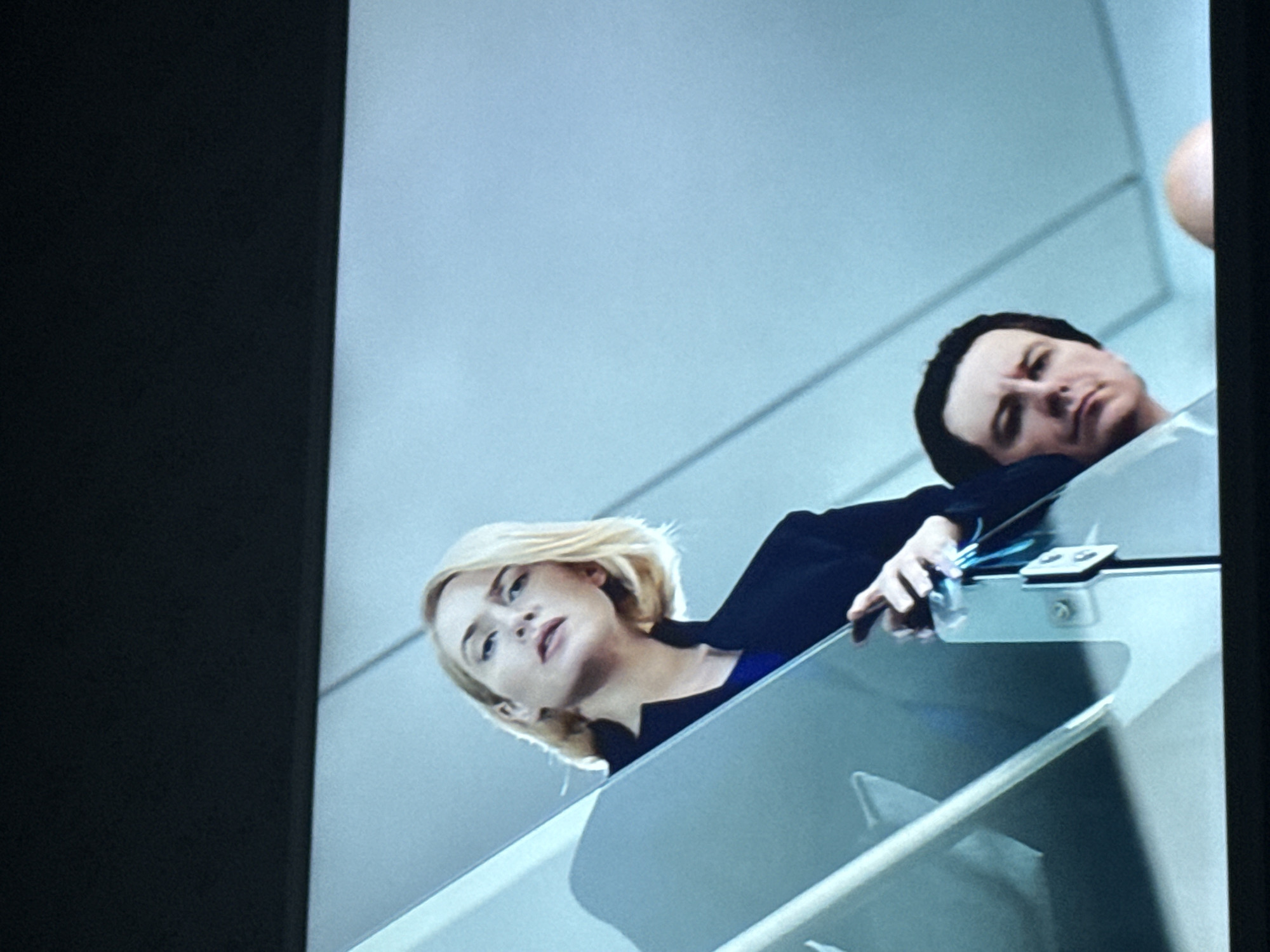 The image shows two individuals, a blonde woman and a dark-haired man, looking down from an elevated position, possibly a balcony or walkway with glass railings. The woman is wearing a dark jacket over a blue top, while the man is wearing a dark jacket as well. They both appear to be focused on something below. The background is a light-colored wall, and there is a partial view of what seems to be another person's head on the right side of the frame. The location of the scene is not discernible from the image alone, therefore, it is not possible to tell if it is Mytishchi, Russia.