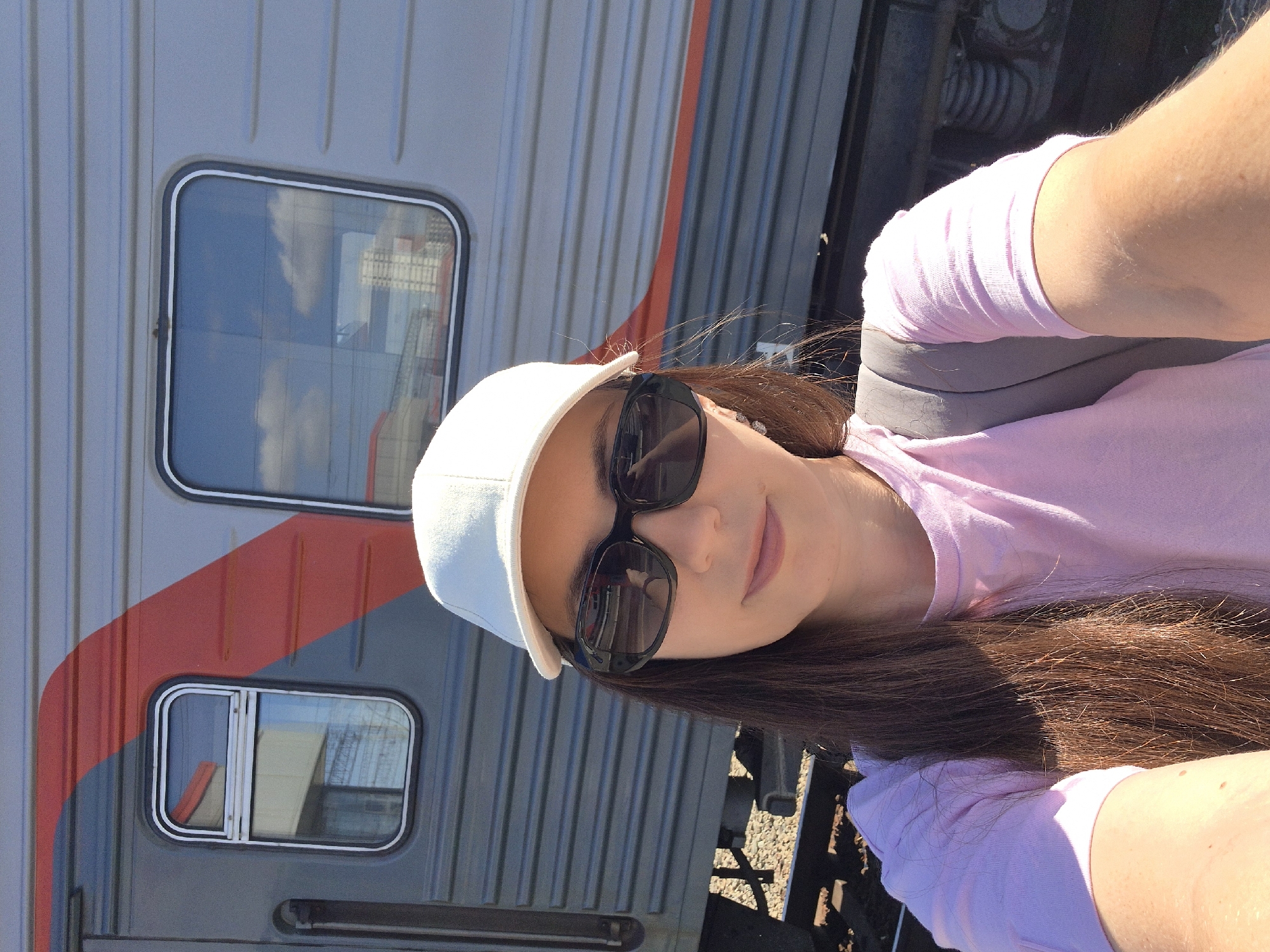 In the provided image, a young woman wearing a white baseball cap and sunglasses takes a selfie in front of a train car. She has long, dark brown hair and is wearing a light pink t-shirt and a gray backpack. The train car is gray with a red stripe and has several windows. The scene appears to be outdoors, possibly at a train station, with gravel visible on the tracks below the train. The reflection in the sunglasses shows another train car. Based on the context of Yekaterinburg, Russia, this photo was likely taken at the Yekaterinburg-Passazhirsky railway station, which is the main railway hub of the city.