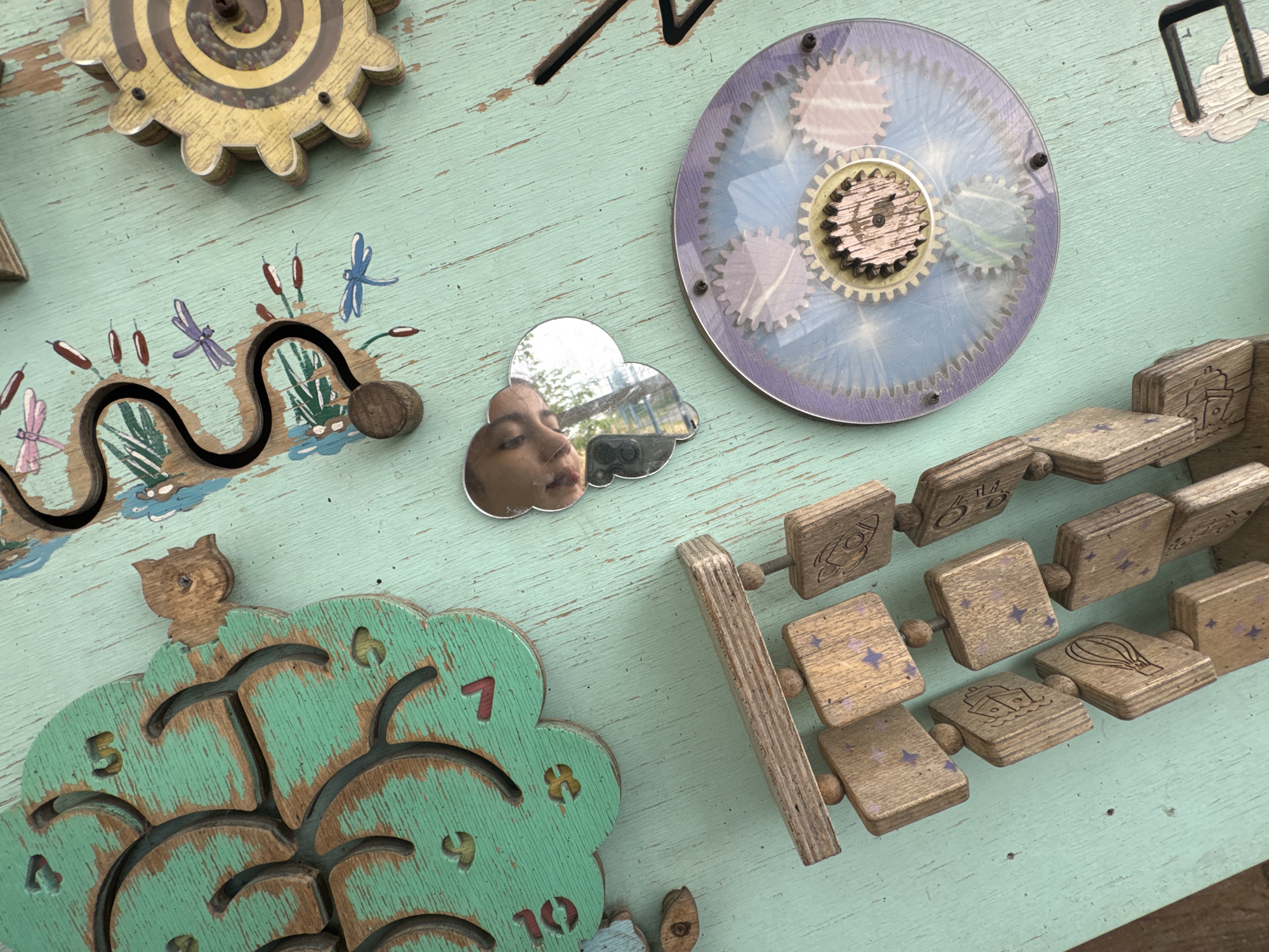 The image is a close-up, overhead shot of a light green, weathered wooden surface with various interactive toy elements attached. The surface appears to be a wall or a large panel, possibly part of a children's play area.
Prominently featured are several wooden toys designed to engage children. In the upper left quadrant, a large, multi-layered wooden gear with a yellow, spiraling maze inside is mounted. Below this, a black, wavy track with small red and blue painted dragonflies and cattails is embedded in the wood, with a small wooden bead running along the track.
To the right of the maze gear, a circular transparent disc with intricate gear designs is affixed. A small, cloud-shaped mirror is placed below this, reflecting a portion of a young child's face and the surrounding environment, which appears to be outdoors with trees and possibly a playground structure.
In the lower half of the image, a large, green, wooden clock face shaped like a tree is visible. The clock face has cut-out numbers (5, 7, 8, 9, 10) and several carved tree branches. A small wooden owl figure sits atop the tree.
To the right of the clock, a chain of wooden blocks is arranged diagonally. These blocks are square and made of natural wood, each featuring a small engraved image such as a rocket, a hot air balloon, a ship, or a star pattern. These blocks appear to be threaded onto a metal rod, allowing them to slide.
The overall scene depicts a well-loved and worn interactive play surface, likely designed for sensory and motor skill development. The location is stated to be in Zaliznychne, Ukraine, although this detail cannot be verified from the image itself. The lighting suggests it is daytime, possibly outdoors due to the reflection in the mirror. There are no people directly visible, only the reflection of a child.