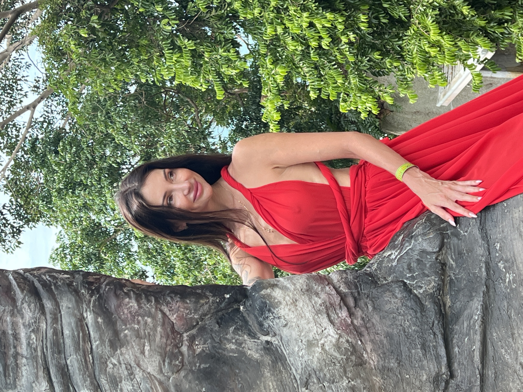 A woman with long dark hair is posing in a red dress on a rocky outcropping in Baan Hua Thanon, Thailand. The woman is smiling, and her left hand is resting on the rock. The background includes trees and some buildings.