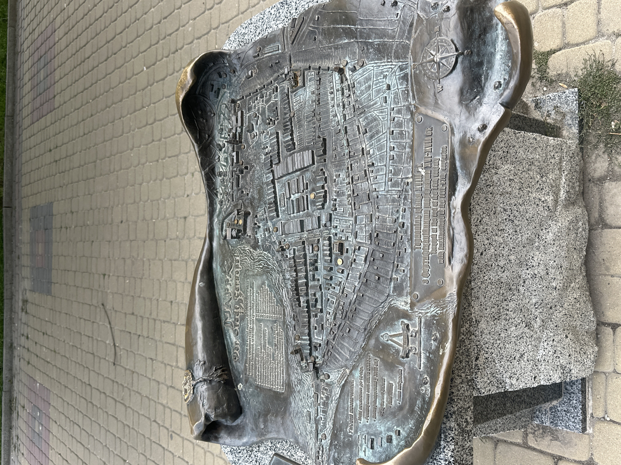 A bronze relief map of Ternopil, Ukraine, is displayed on a stone pedestal. The map depicts the city in a historical style, with raised buildings and streets. Text in Ukrainian is inscribed on the map, providing information about the city's history and landmarks. A compass rose is visible in the lower right corner of the map. The map is located outdoors on a paved walkway. The weather appears to be overcast.