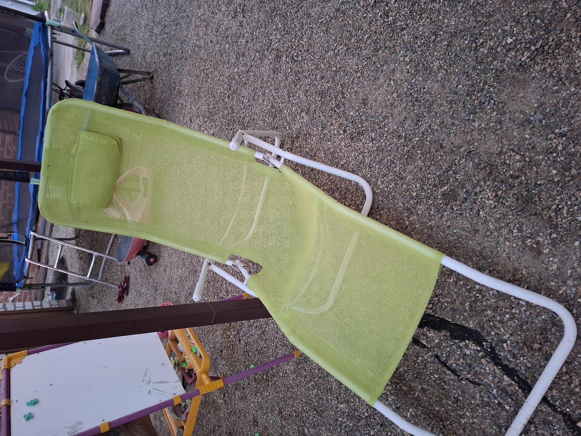 A green mesh lounge chair with a white metal frame sits on a gravel surface. The chair is positioned in a backyard in Korenovsk, Russia. In the background, a trampoline with a blue mat is visible, as well as a wheelbarrow. To the left of the chair is a purple and yellow easel with a whiteboard. The scene appears to be outdoors, possibly in a residential area.