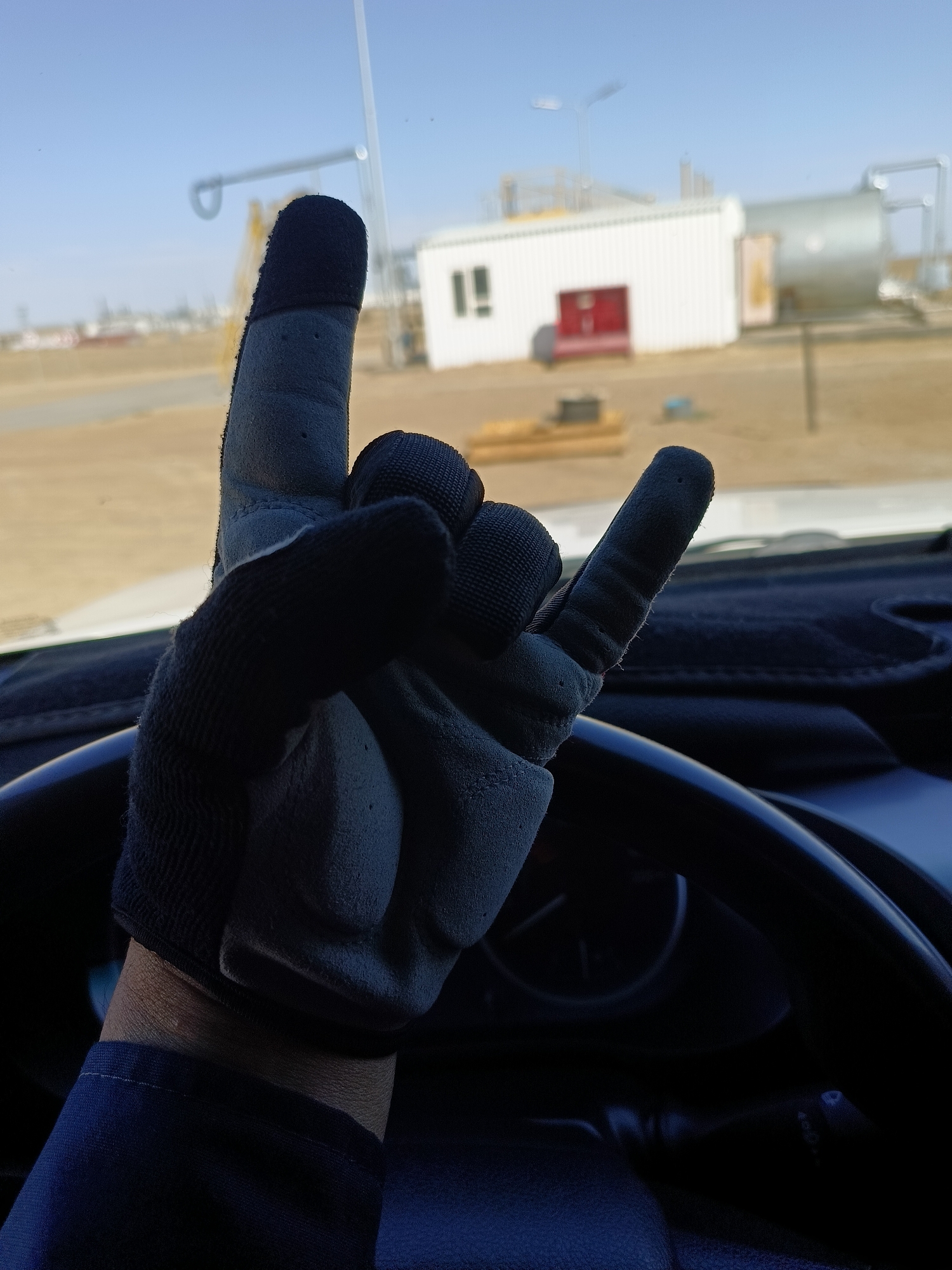 A gloved hand is held up in front of a steering wheel, forming the rock on gesture. The background is blurred and shows an industrial area with a white building and a large silver tank under a clear blue sky. The scene appears to be outdoors in a dry, arid environment, possibly within an industrial facility or construction site. The image is taken from the driver's perspective inside a vehicle.