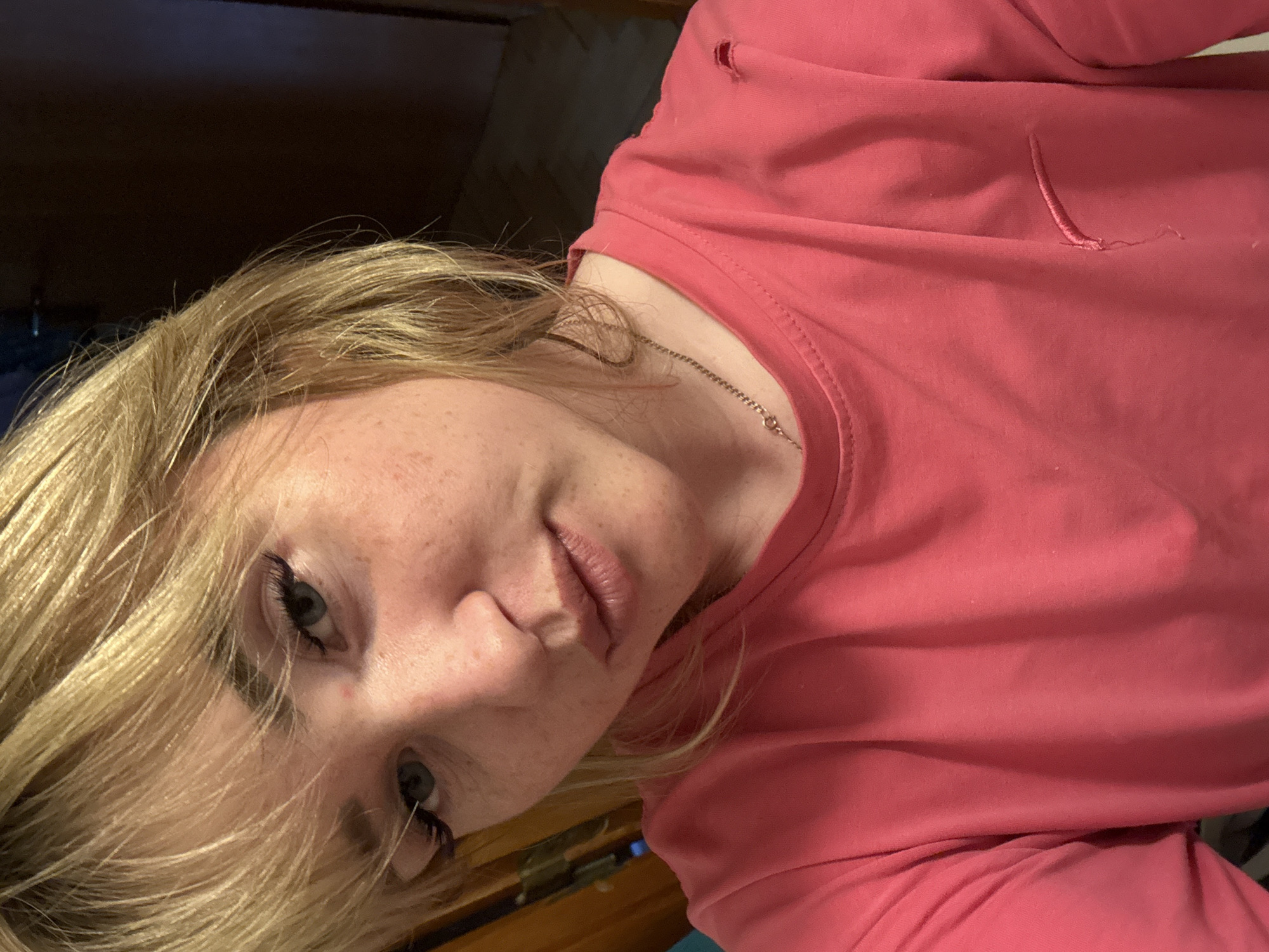 This media file is a portrait of a young person with blonde hair, freckles, and blue eyes, wearing a pink long-sleeved shirt. The person is looking towards the camera with a neutral expression. The image is close-up, focusing on the person's face and upper torso. The background is dark and out of focus, making the subject the central element.
There are two notable details on the shirt: a small hole near the shoulder and a loose thread on the chest area, suggesting the garment might be old or slightly damaged. A thin silver necklace is visible around the person's neck. The lighting appears to be indoor, possibly artificial, casting shadows that highlight the contours of the face. The overall mood is subdued and intimate due to the close framing and gentle lighting. The image appears to be a selfie or a personal photograph taken in a casual setting, likely at home. There is no visible text in the image. The location context is provided as Марковский, Russia, but there are no specific visual cues within the image to confirm this location.